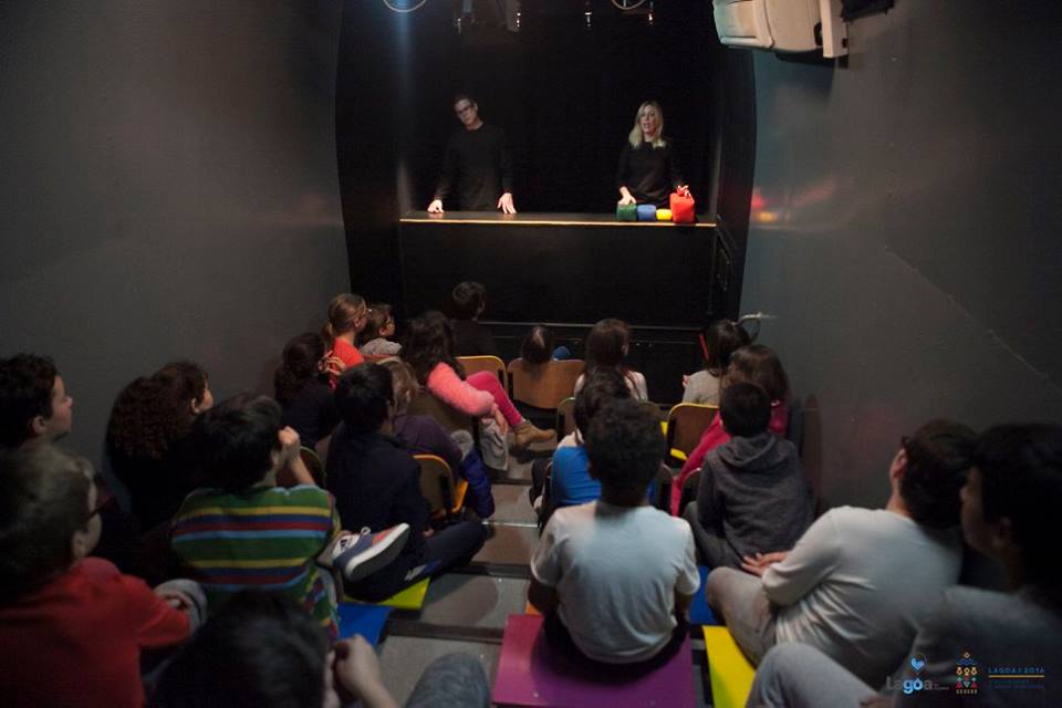 Crianças sentadas em arquibancada assistindo a uma apresentação de teatro de fantoches com dois animadores no palco.
