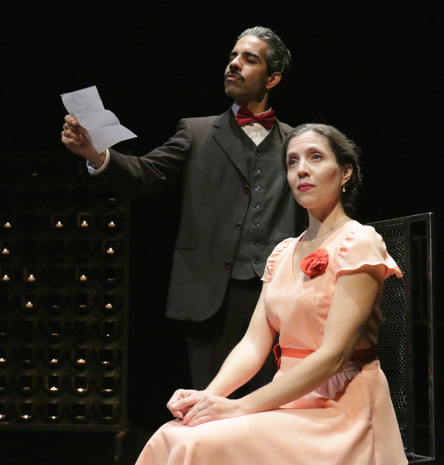 Homem em fato preto com gravata borboleta vermelha a segurar um papel, mulher sentada de vestido cor de salmão com flor vermelha, ambos em cenário de teatro.