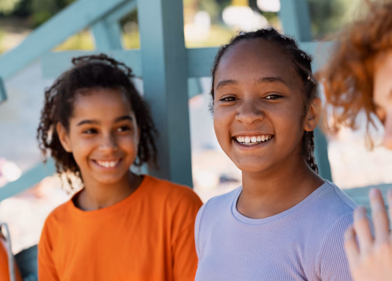 Two smiling girls with braided hair, one wearing a lavender shirt and the other an orange shirt, outdoors near a light blue structure.