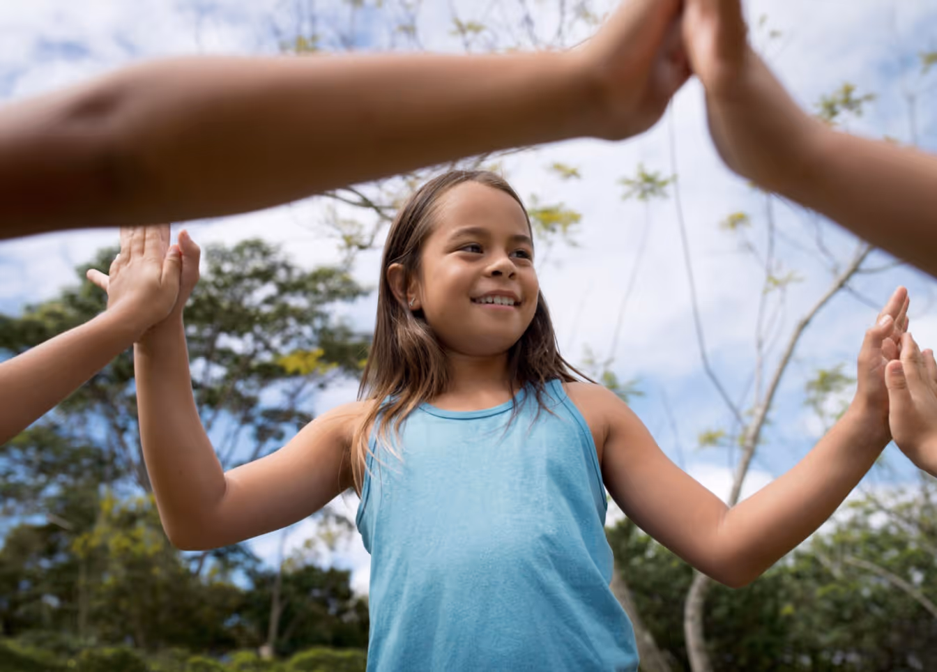 Smiling girl in blue tank top outdoors giving high fives with other children.