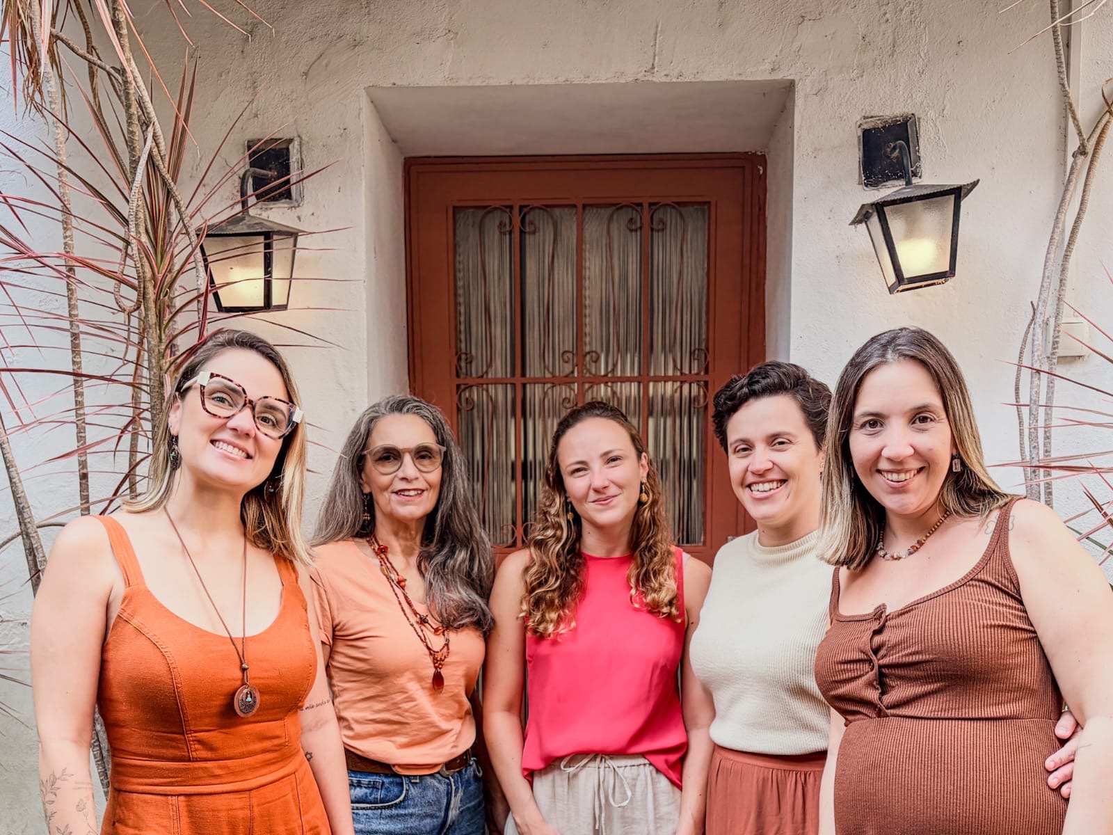 Five women standing close together smiling outdoors in front of a closed door and wall lamps.