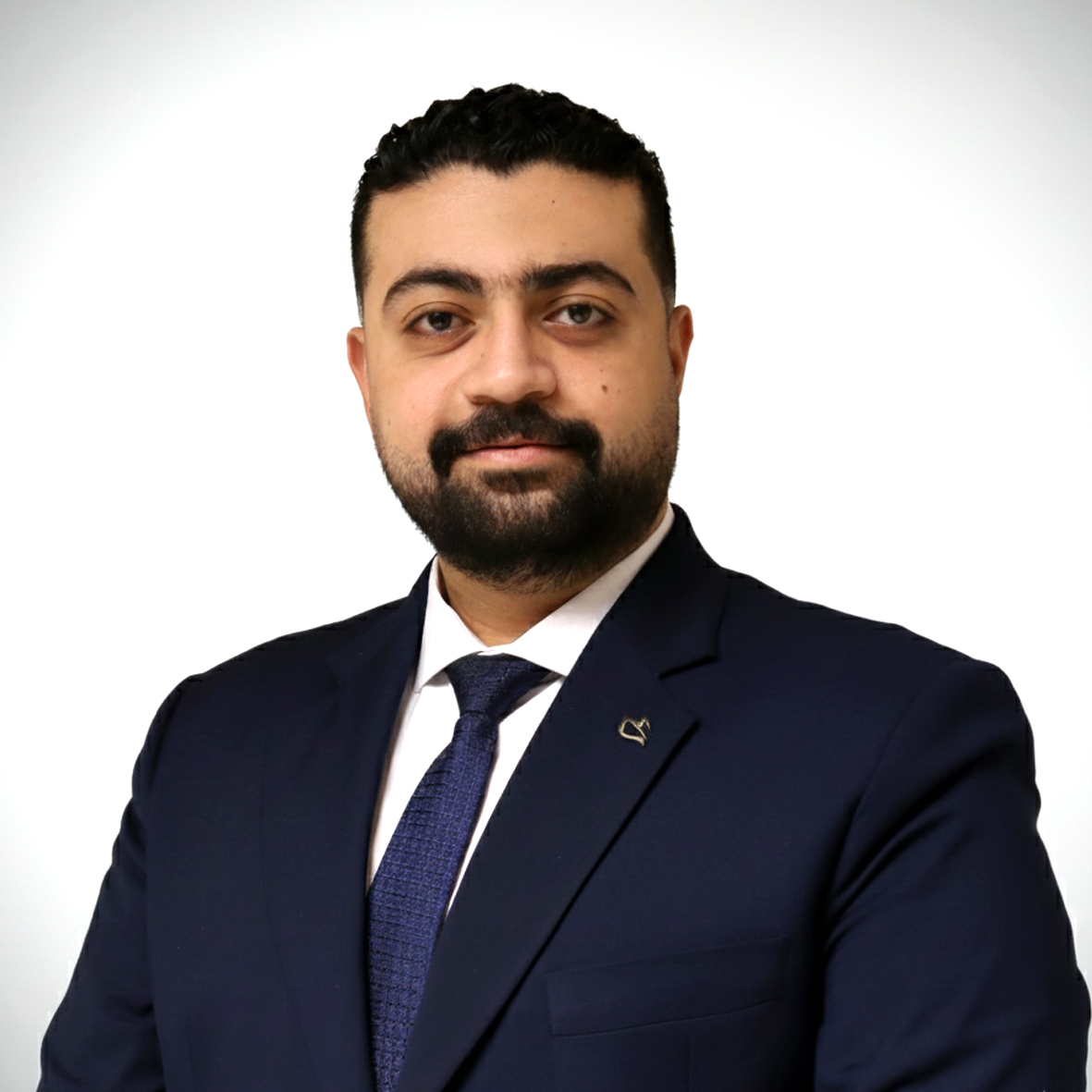 Man with black hair and beard wearing a navy blue suit, white shirt, and blue tie against a white background.