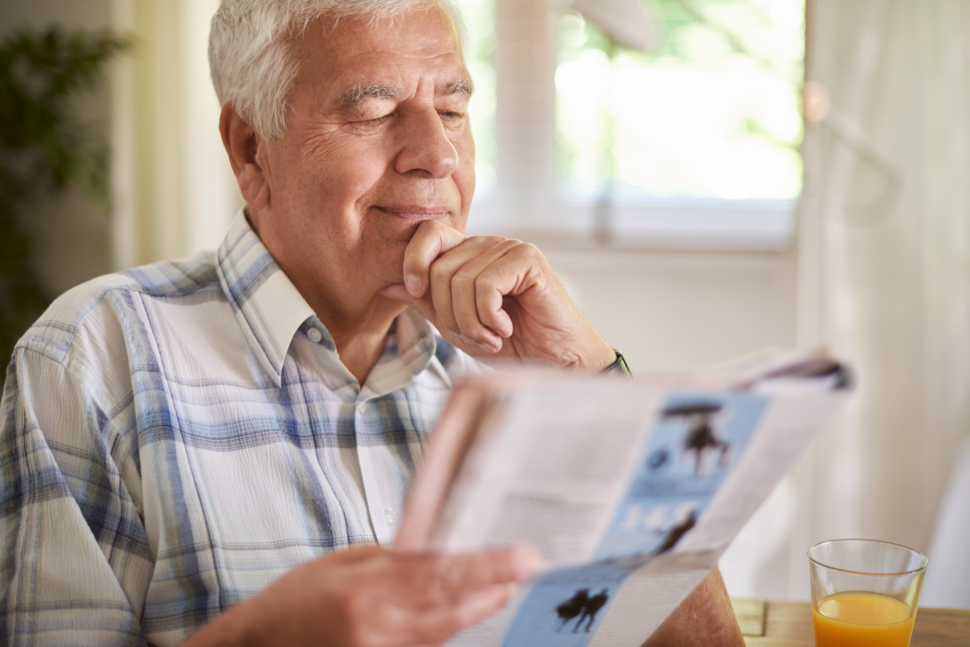 Elderly man reading a newspaper with a glass of orange juice on the table beside him.