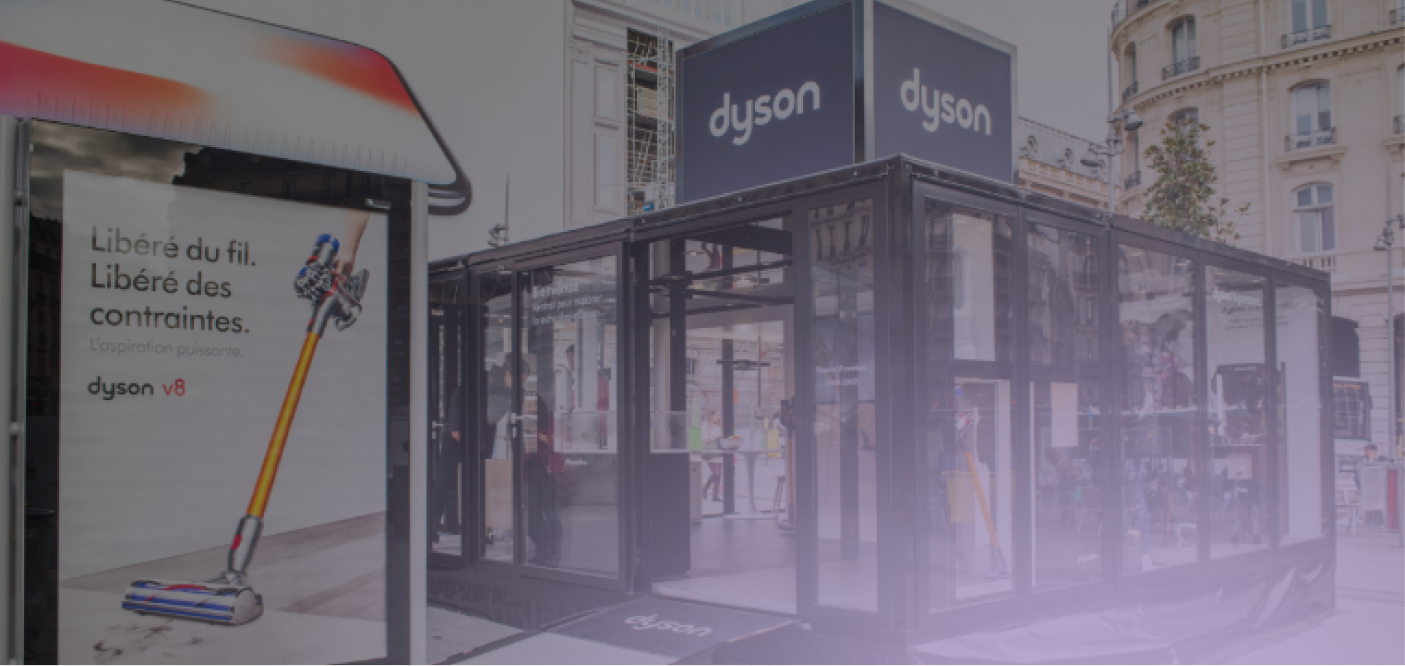 Outdoor Dyson showroom with transparent walls displaying vacuum cleaners and an advertisement for Dyson V8 cordless vacuum in French.