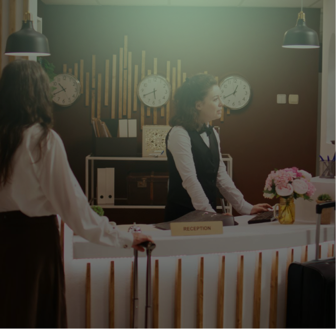 Hotel receptionist in uniform assisting a woman guest standing with a suitcase at the front desk with clocks showing different time zones behind.