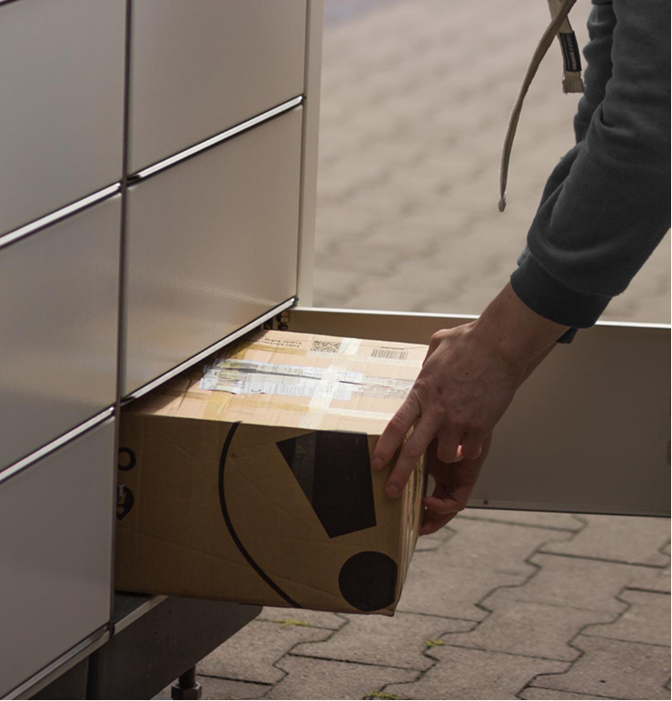 Person retrieving a taped cardboard package from a metal parcel locker outdoors on a paved surface.