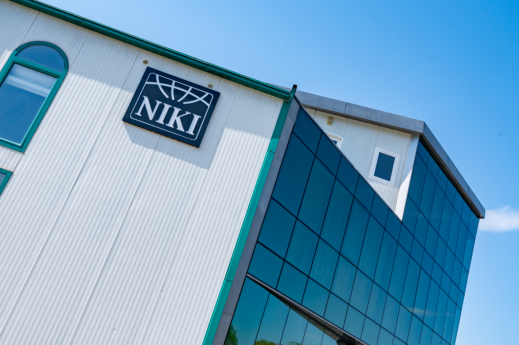 Green two-seater helicopter parked on a concrete surface in front of a building with plants and trash bins nearby.