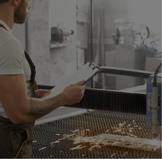 Man with tattooed arm holding clipboard while supervising metal cutting with sparks flying in an industrial workshop.