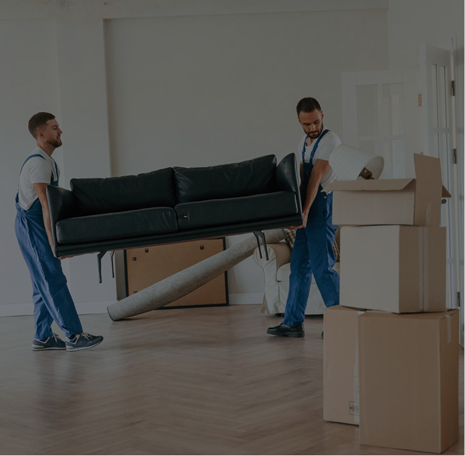 Two men in overalls carrying a dark green sofa in a bright, empty room with stacked cardboard boxes nearby.