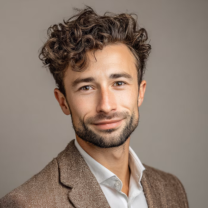 Man with curly brown hair and beard wearing a brown blazer and white shirt, smiling against a plain background.
