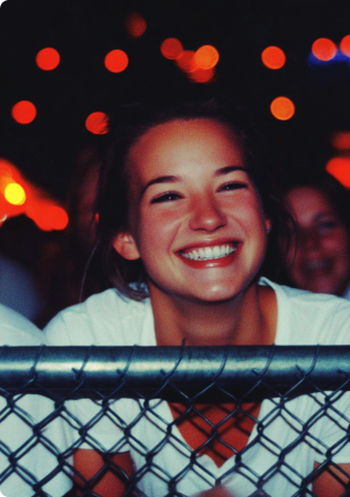 Smiling young woman leaning on a metal fence with blurred colorful lights in the background.