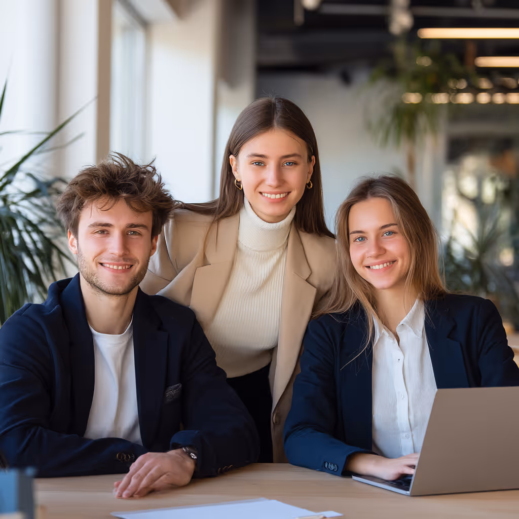Three young professionals smiling and posing at a desk with a laptop in a modern office.