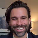 Smiling man with dark hair and beard in front of a window showing city buildings.