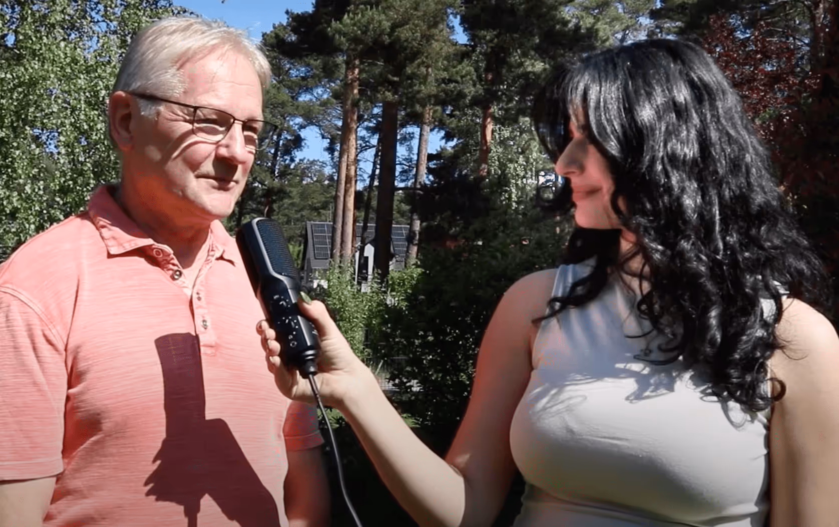 Eine Frau mit langen schwarzen Haaren hält ein Mikrofon vor einen älteren Mann mit Brille und rosa Polohemd in einem sonnigen Park.