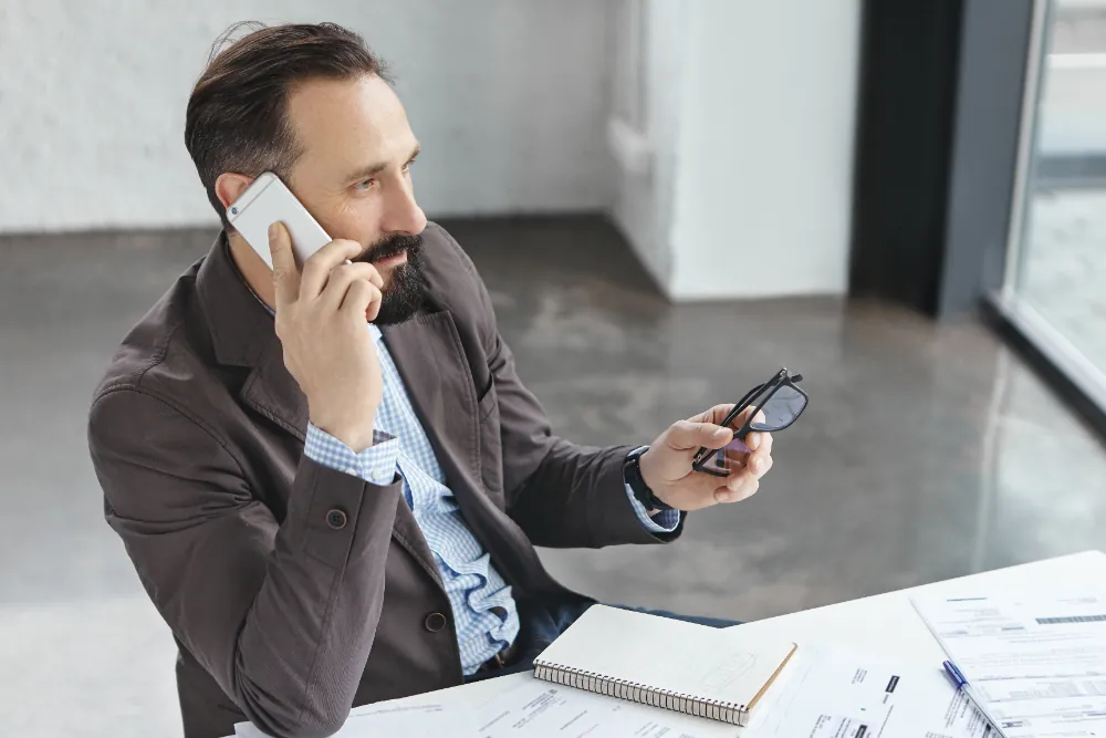 Ein Mann mit Bart hält eine Brille in der Hand und spricht am Telefon, während er an einem Tisch mit Dokumenten sitzt.