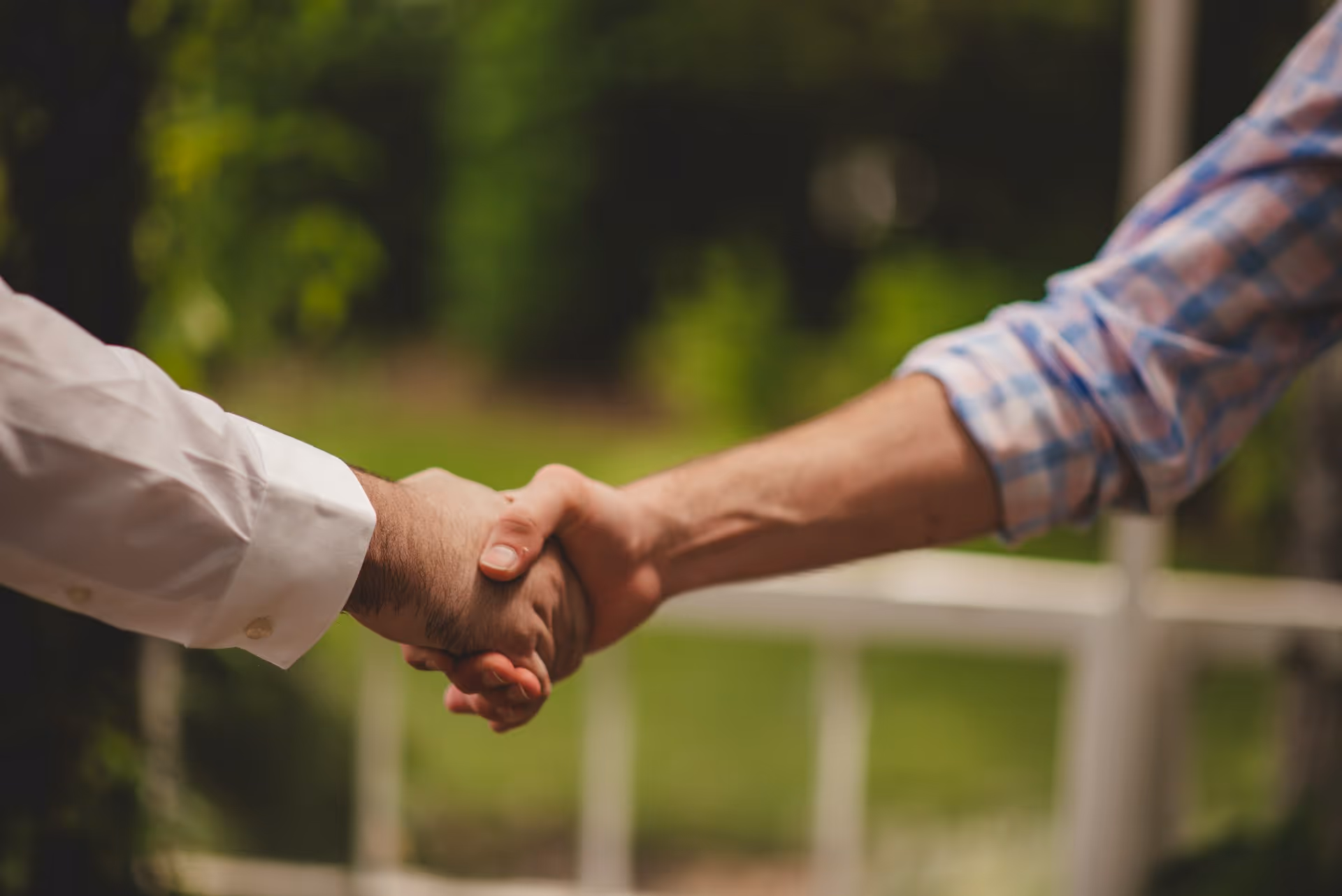 Zwei Personen geben sich im Freien die Hand vor unscharfem Hintergrund aus Grünflächen und Gartenzaun.