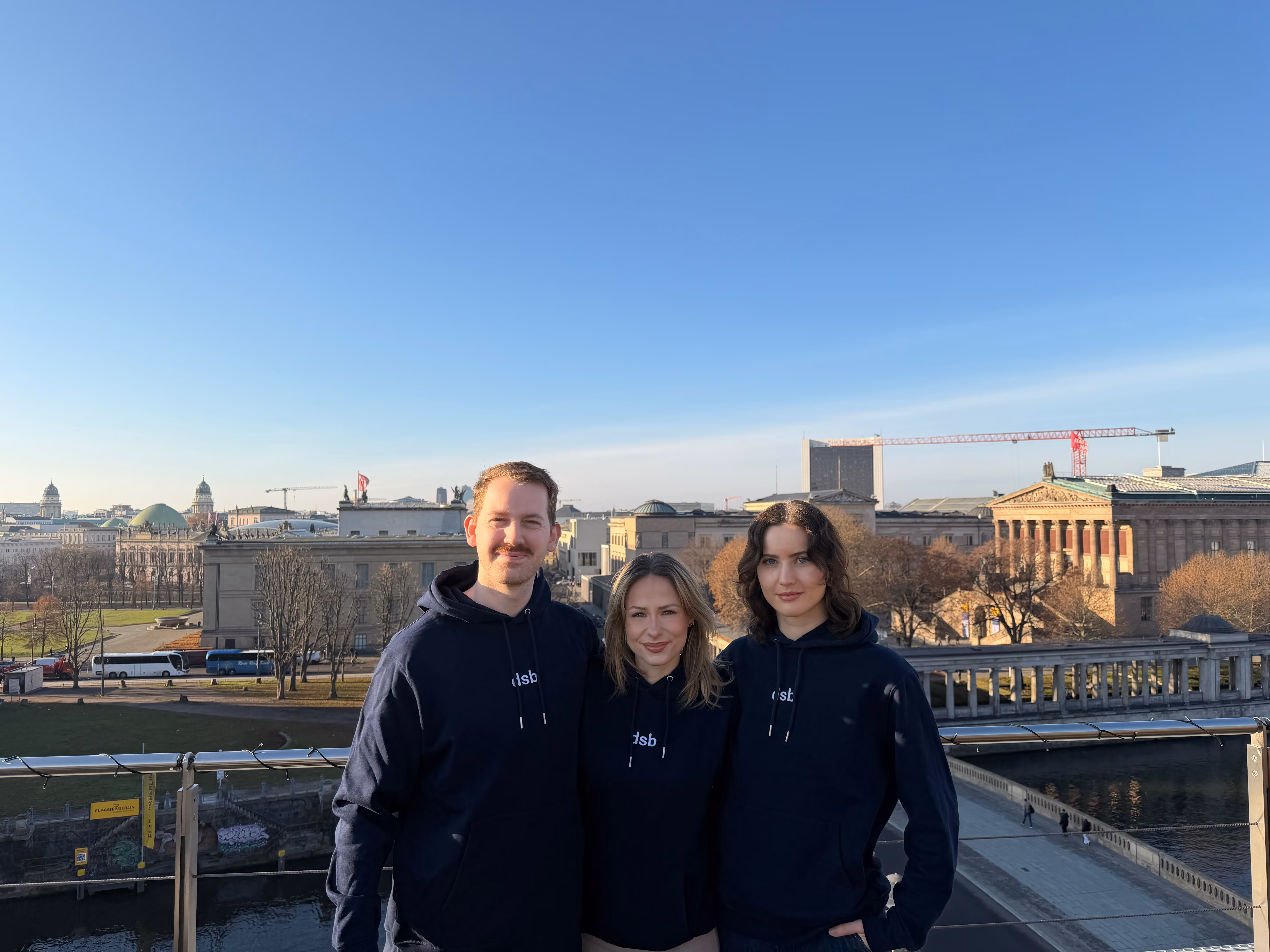 Drei Personen tragen dunkelblaue Kapuzenpullover mit weißem Logo und posieren vor einer Brücke mit historischen Gebäuden im Hintergrund bei klarem Himmel.