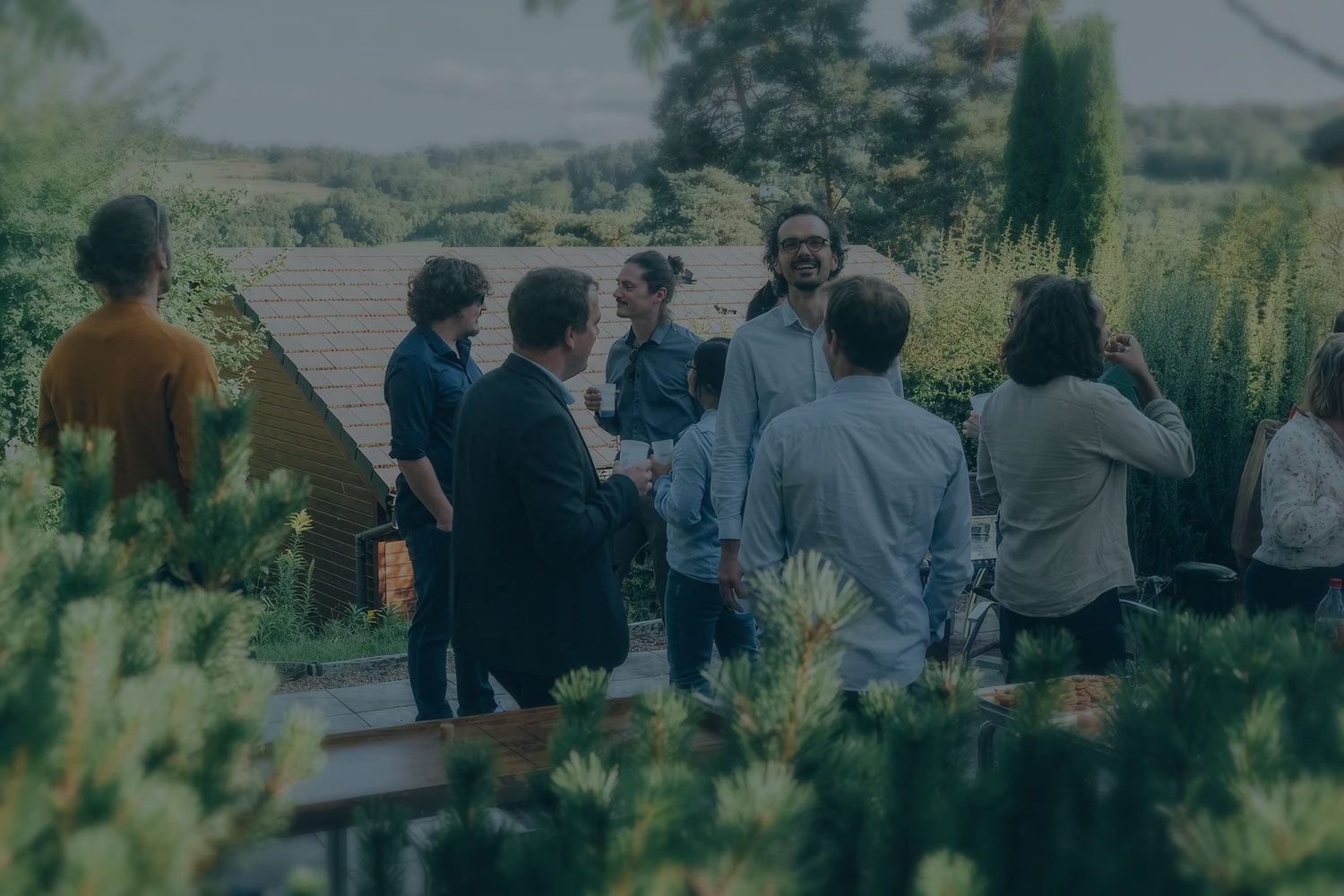Un groupe de personnes discutant en plein air lors d'un rassemblement, avec des arbres et une cabane en arrière-plan.