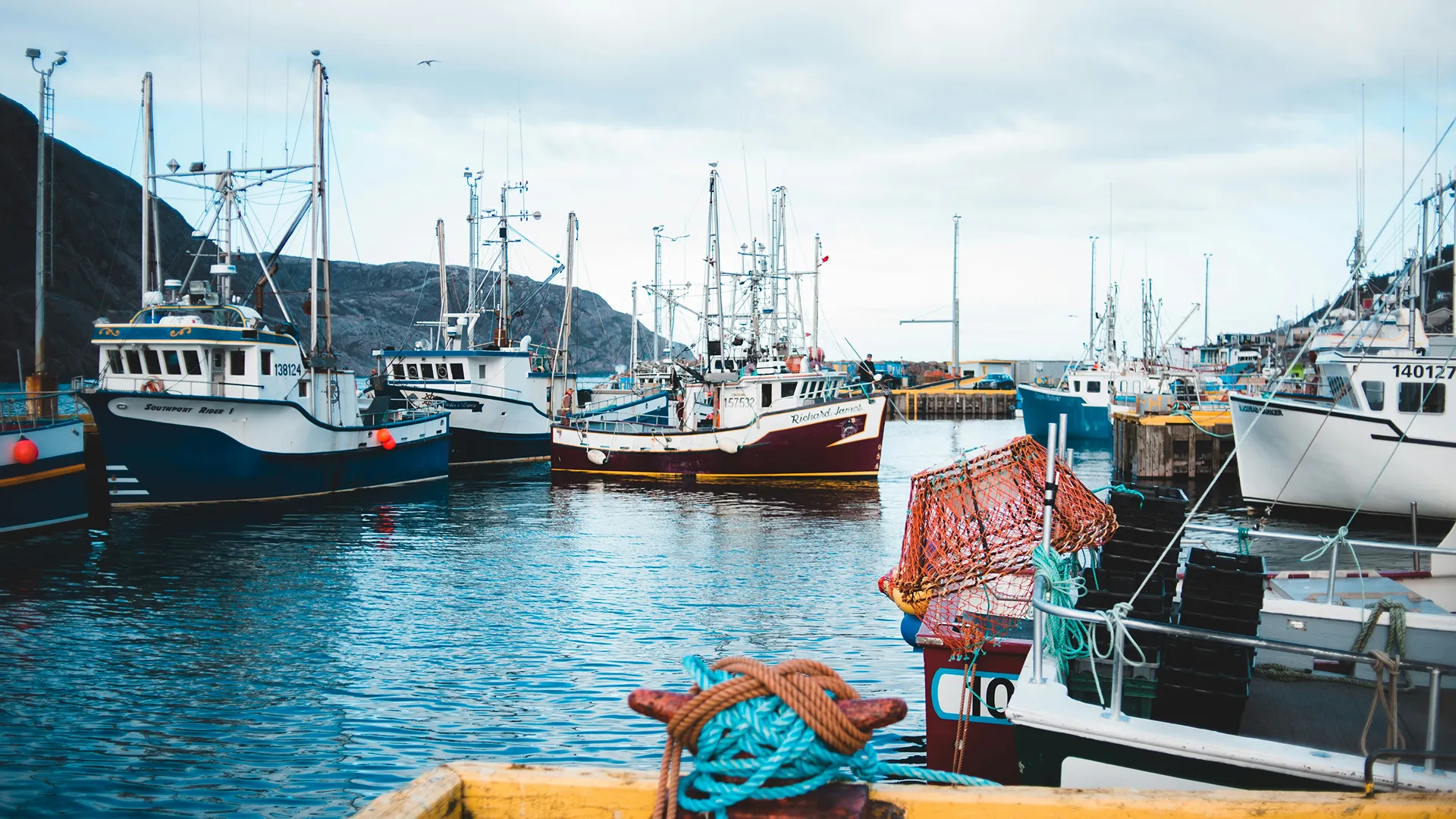 Bateaux de pêche amarrés dans un port calme avec des montagnes en arrière-plan et un filet orange posé sur le quai.