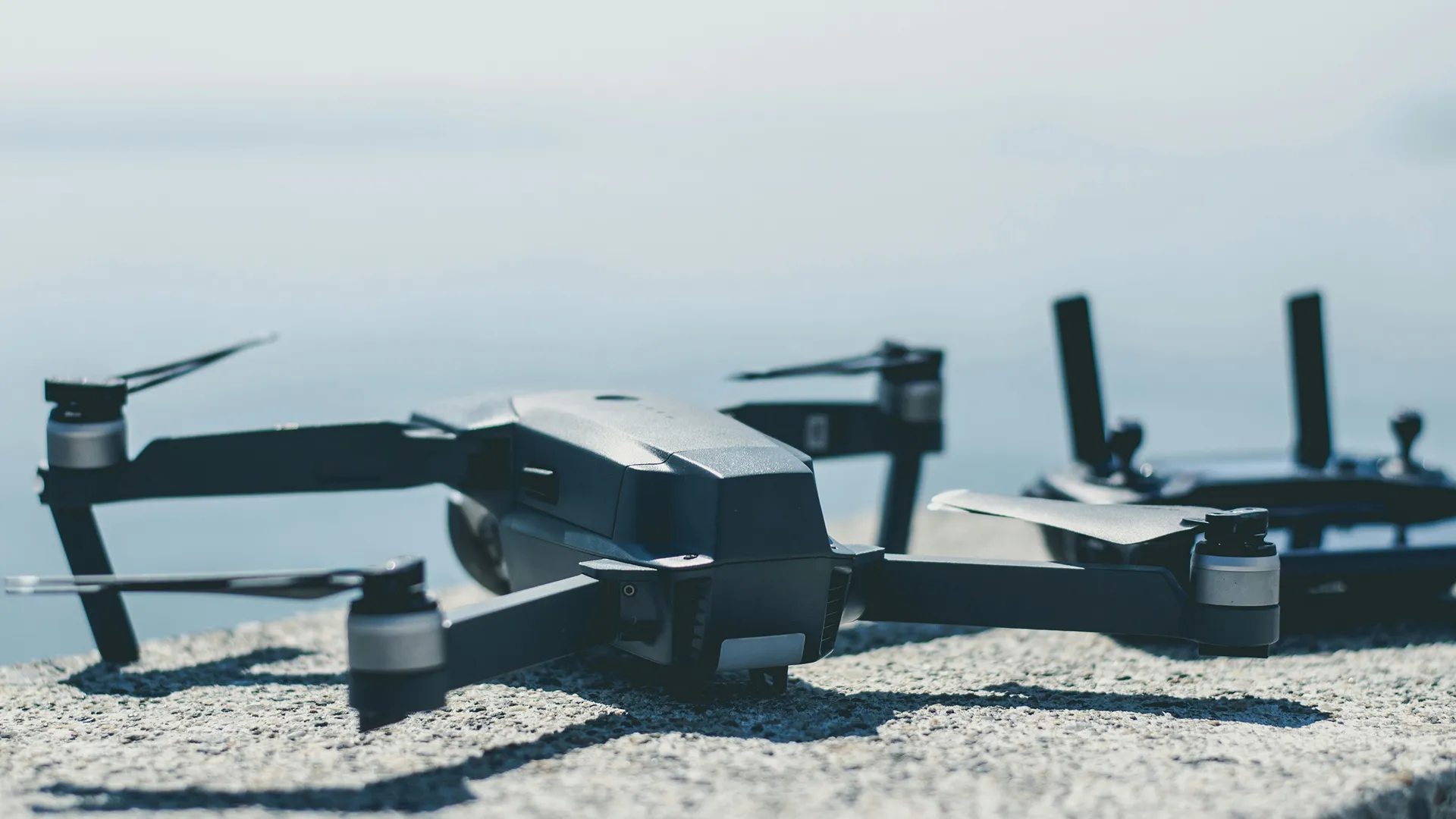 Black drone sitting on a flat surface with a remote control out of focus in the background.