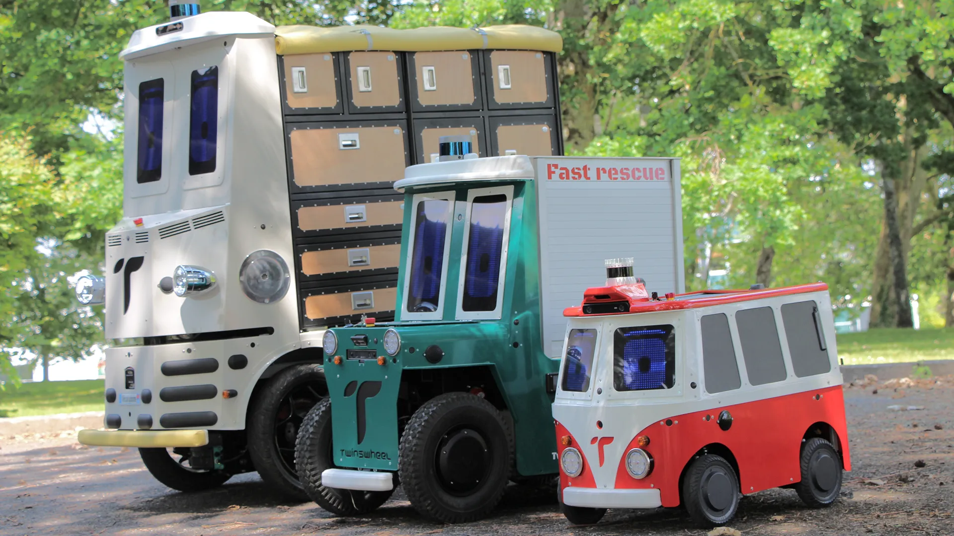 Three small autonomous vehicles of different colors parked on a path with green trees in the background.