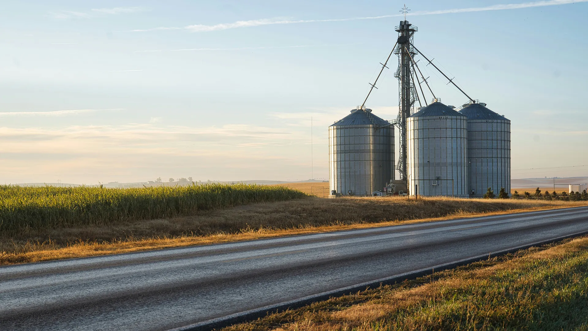 Trois silos métalliques agricoles à côté d'un champ et d'une route sous un ciel clair au coucher du soleil.