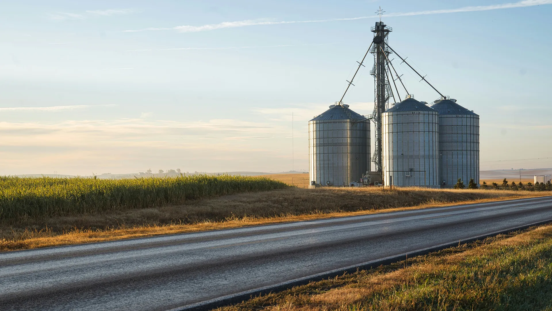Three metal grain silos near a rural road lined with crops at sunset.