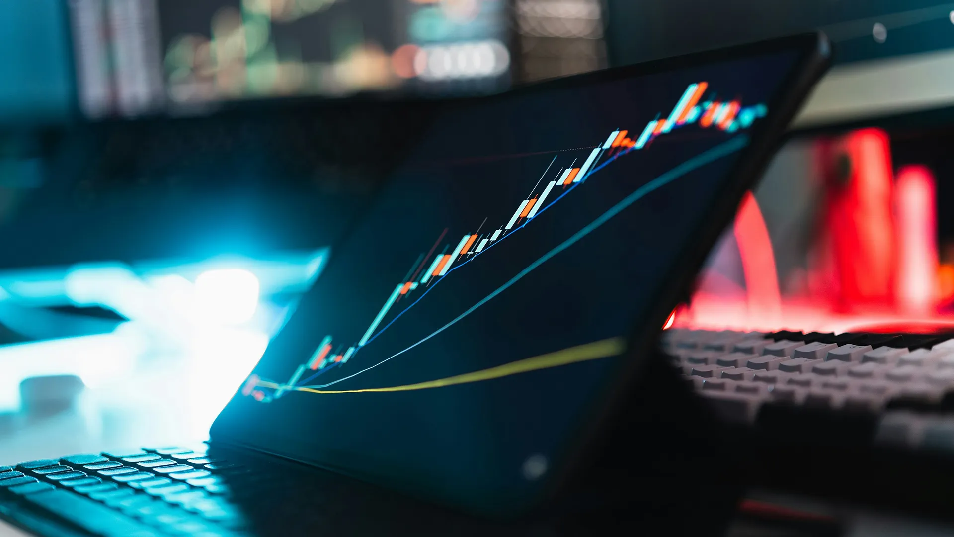 Laptop showing a rising financial chart with candlesticks, surrounded by a keyboard and a blurred screen in the background.