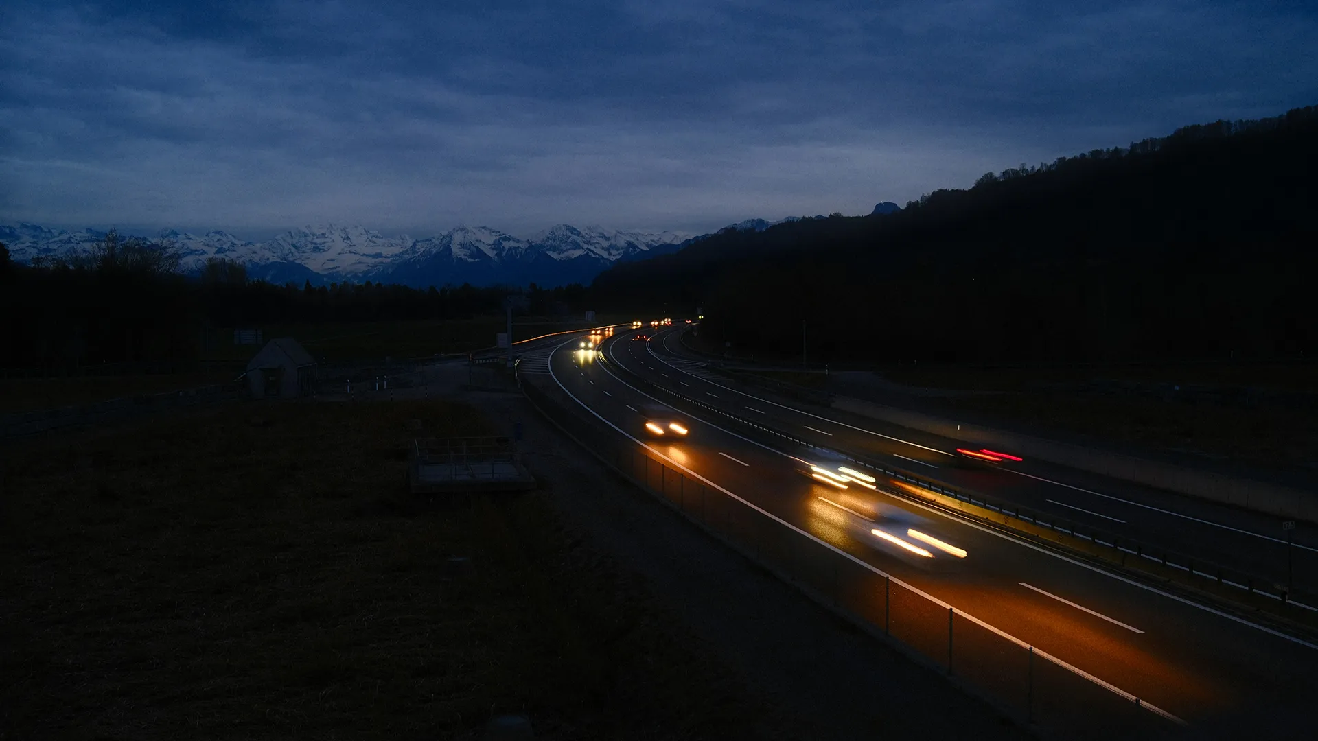 Route sinueuse avec des phares de voitures allumés la nuit, montagnes enneigées à l'arrière-plan.