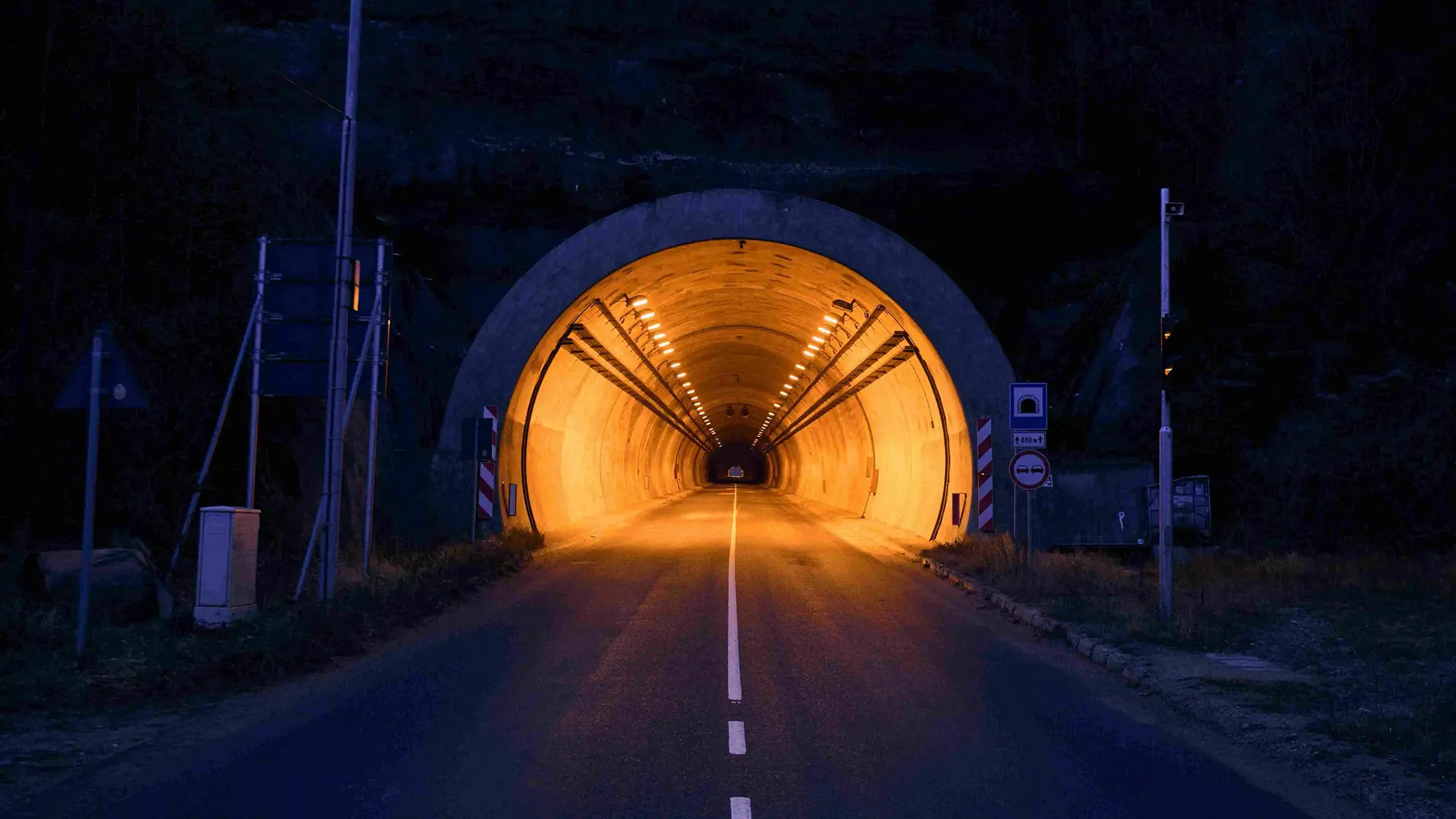 Road tunnel illuminated at night with a two-way road and traffic signs at the entrance.