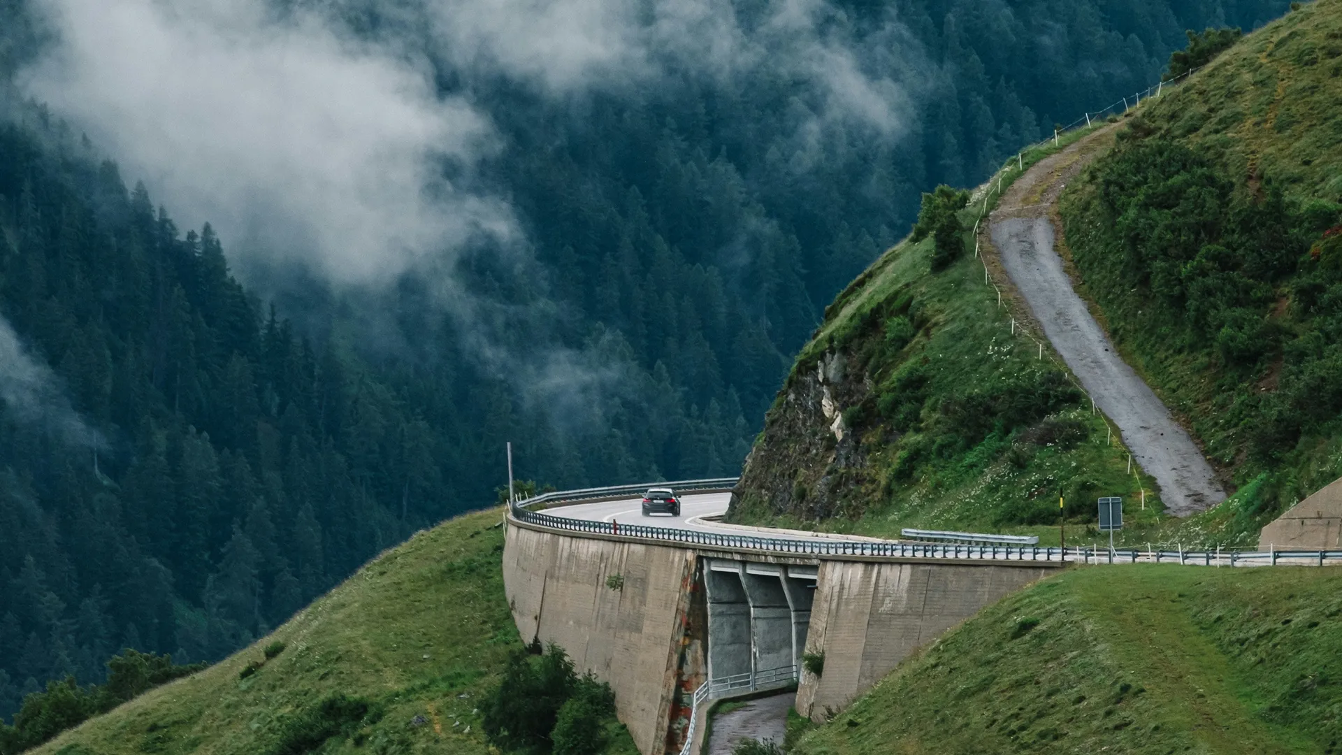 Black car driving on a winding mountain road over a wooded valley in the fog.