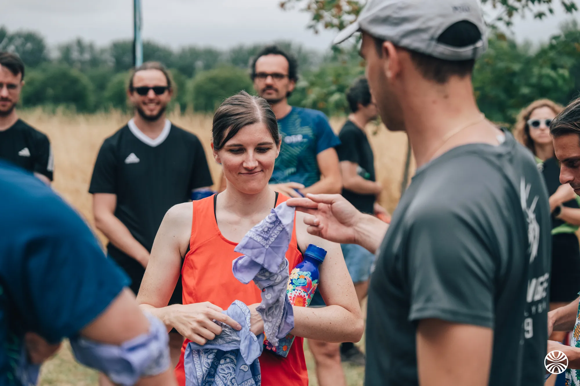 Un groupe de personnes en tenue de sport en plein air, une femme en débardeur orange tient un foulard violet et une bouteille d'eau bleue ornée de fleurs, un homme en gris lui tend un foulard.