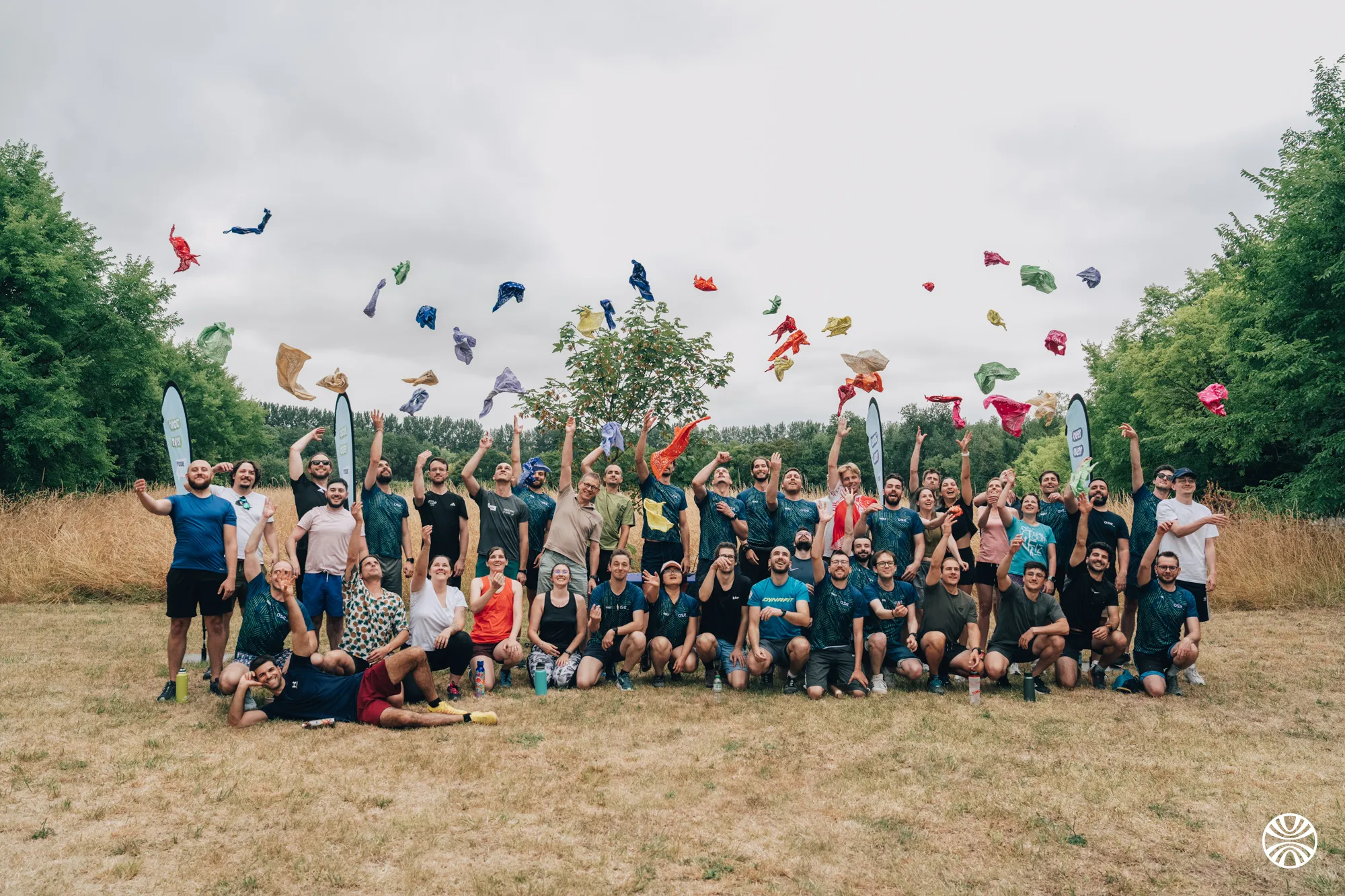 Un grand groupe de personnes souriantes, en tenue sportive, lance des foulards colorés en l'air dans un champ avec des arbres en arrière-plan.