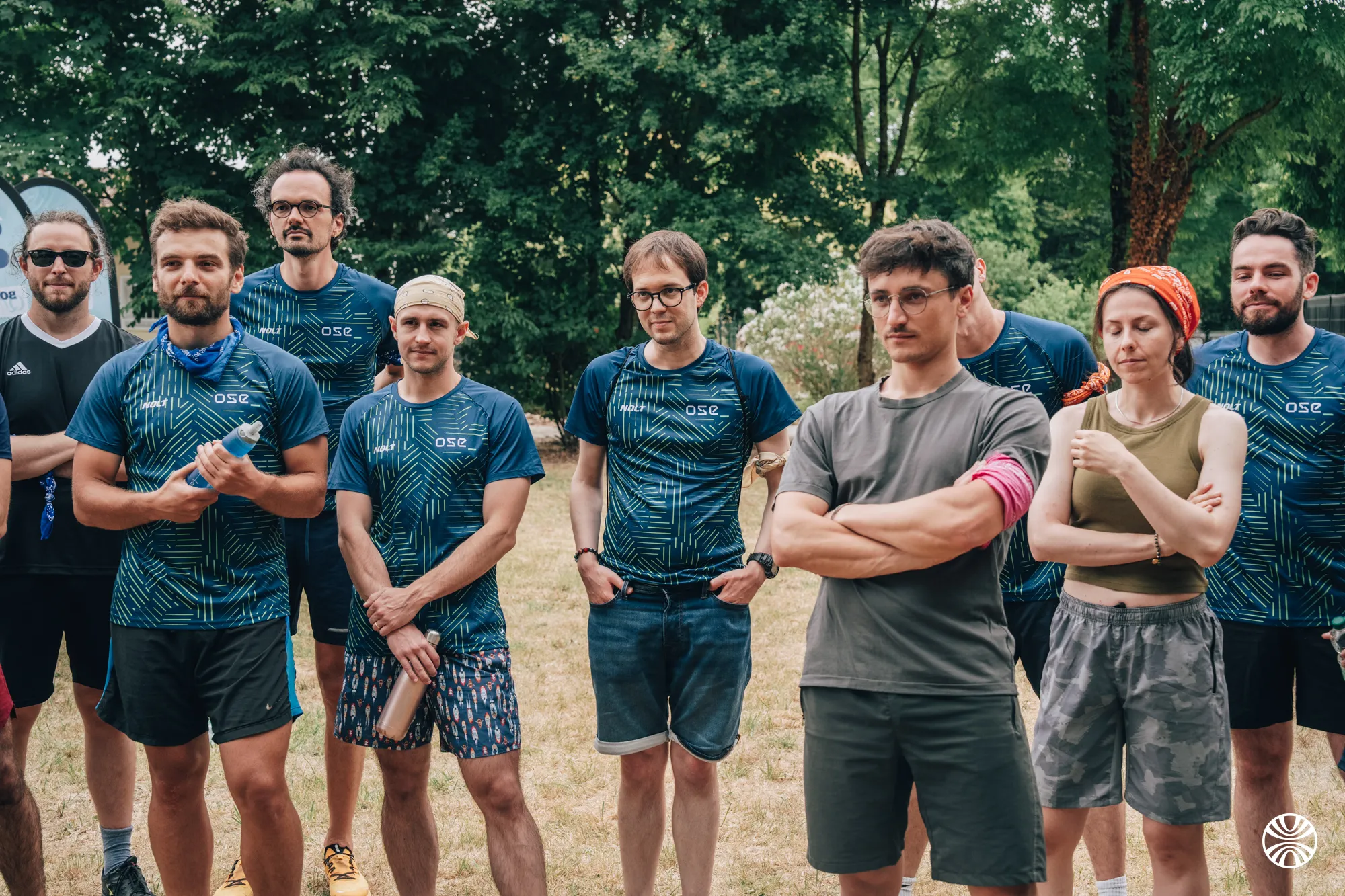 Un groupe de neuf jeunes adultes en tenue de sport posant dehors sur une pelouse avec des arbres en arrière-plan.