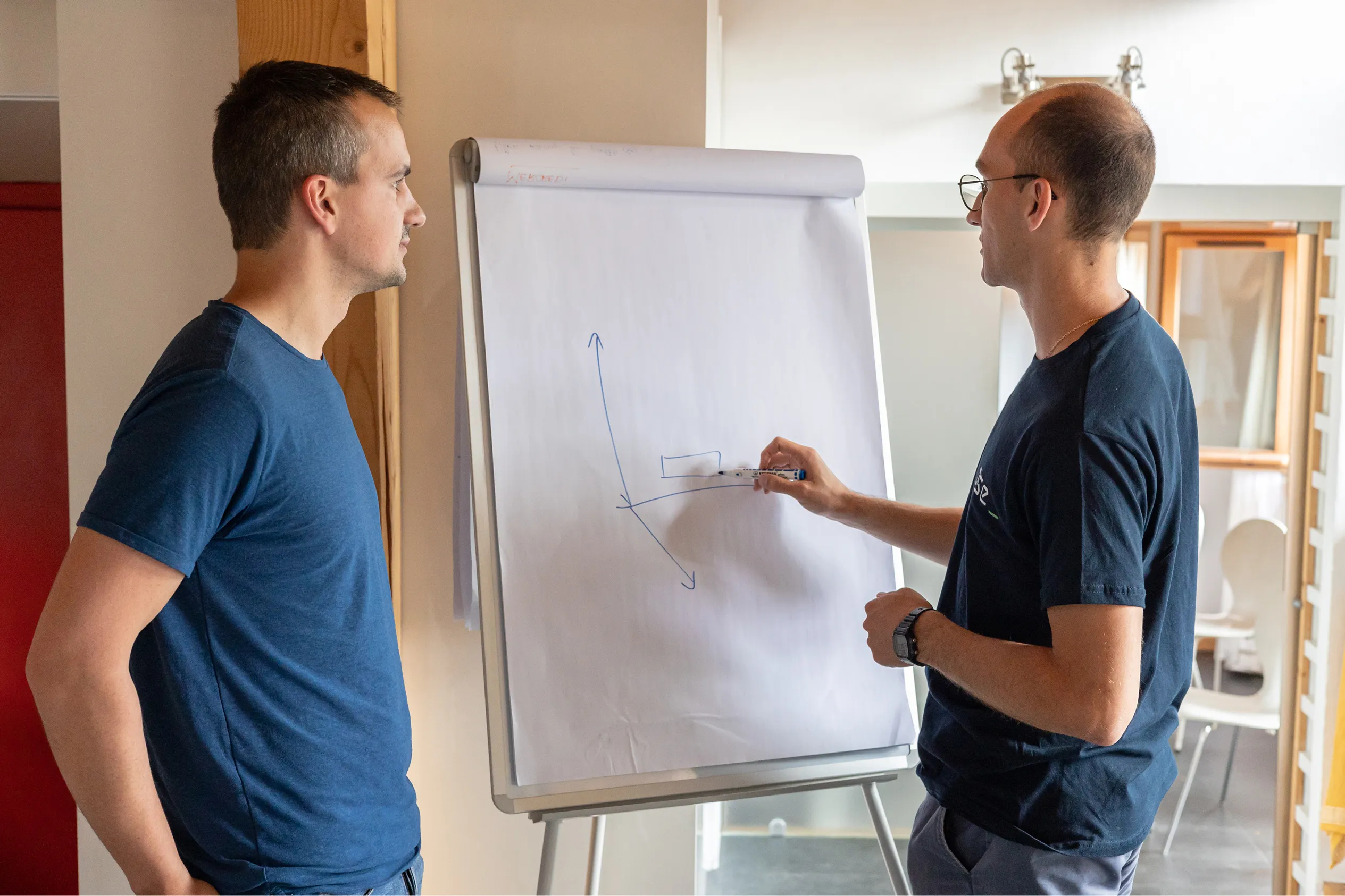 Deux hommes discutent devant un tableau blanc où l'un dessine un graphique avec des flèches sur un papier.