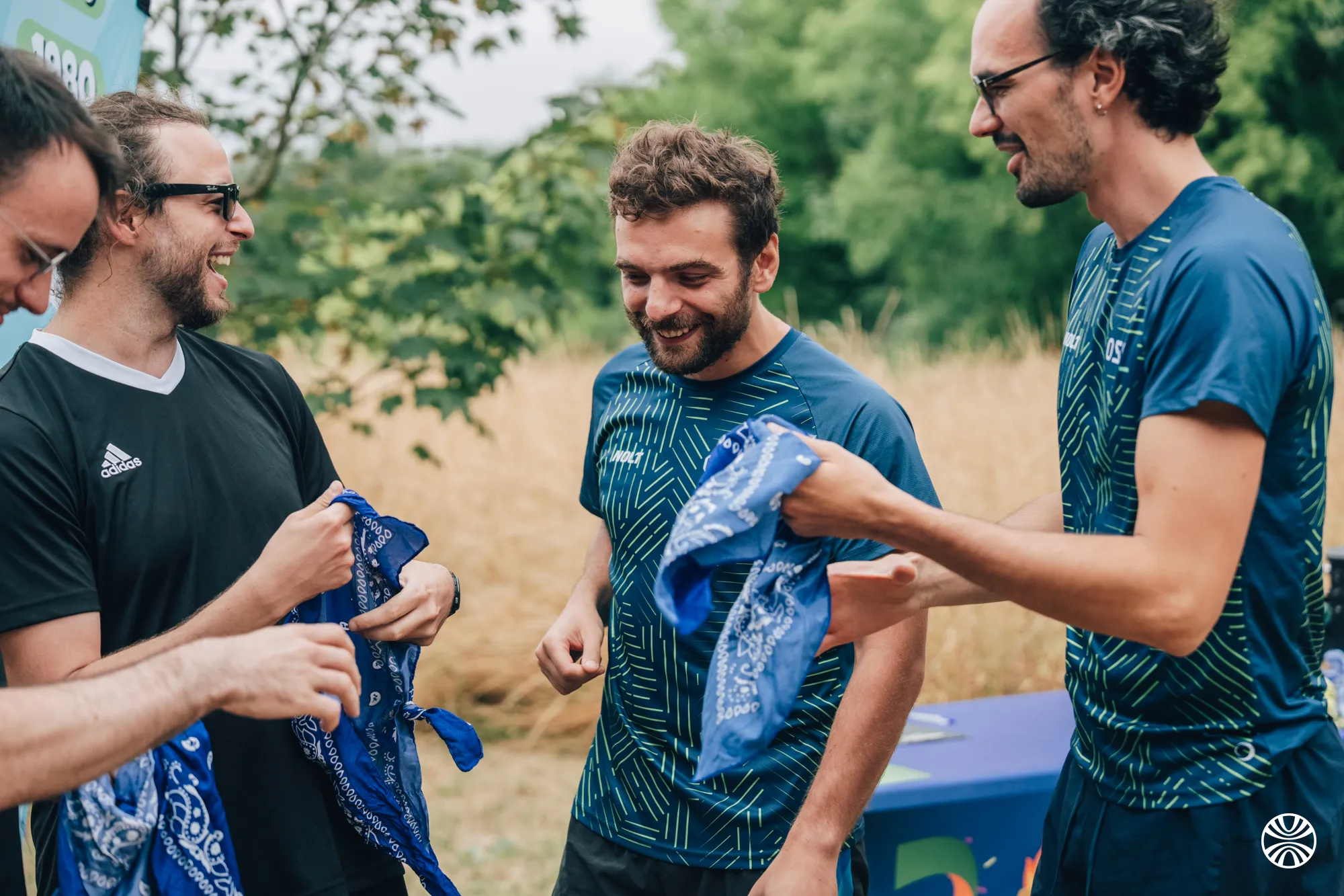 Quatre hommes souriants en tenue sportive tenant des bandanas bleus à l'extérieur près d'une table.