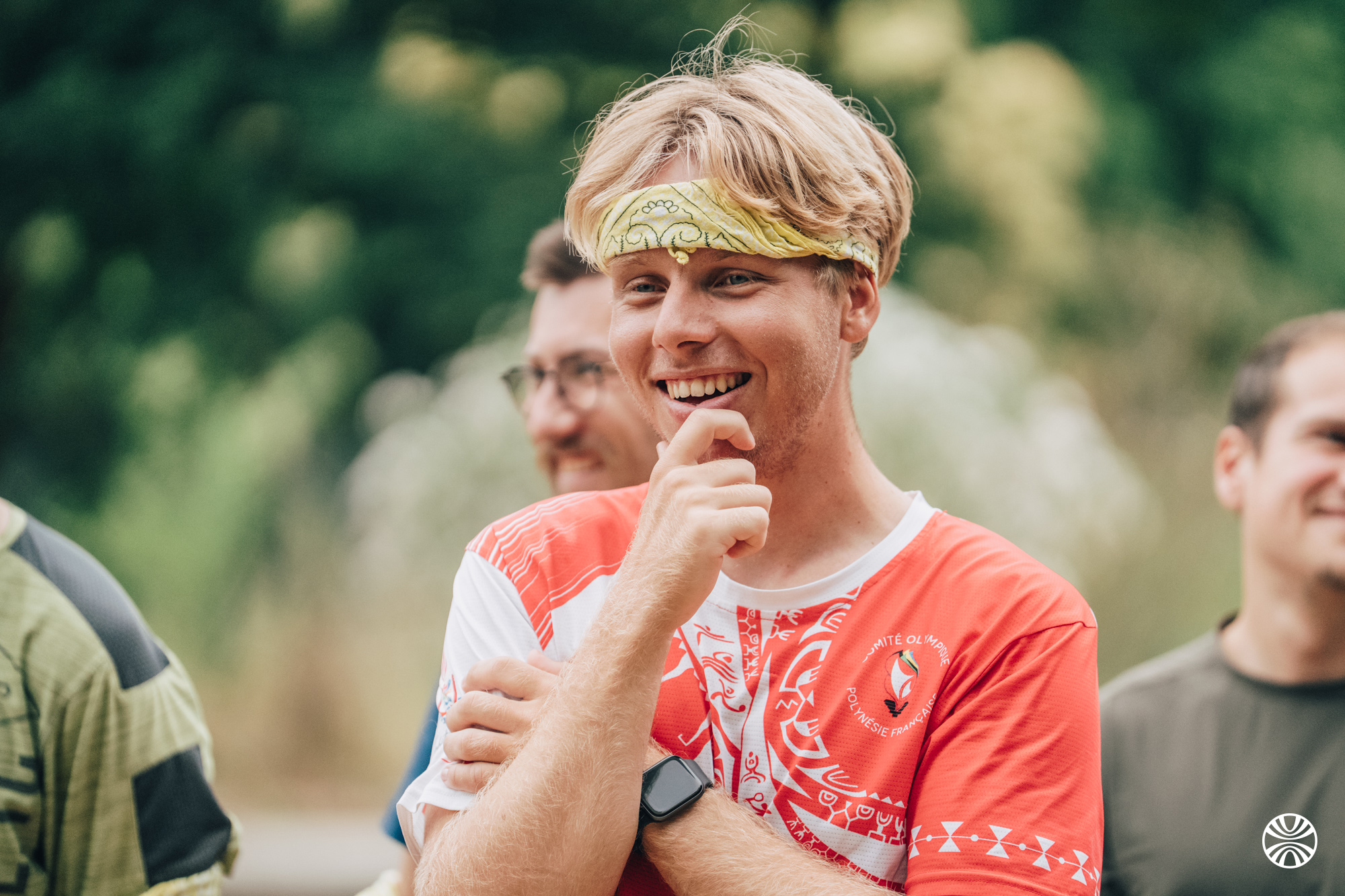 Quatre hommes discutant à l'extérieur dans un environnement verdoyant avec un lac et des collines en arrière-plan.