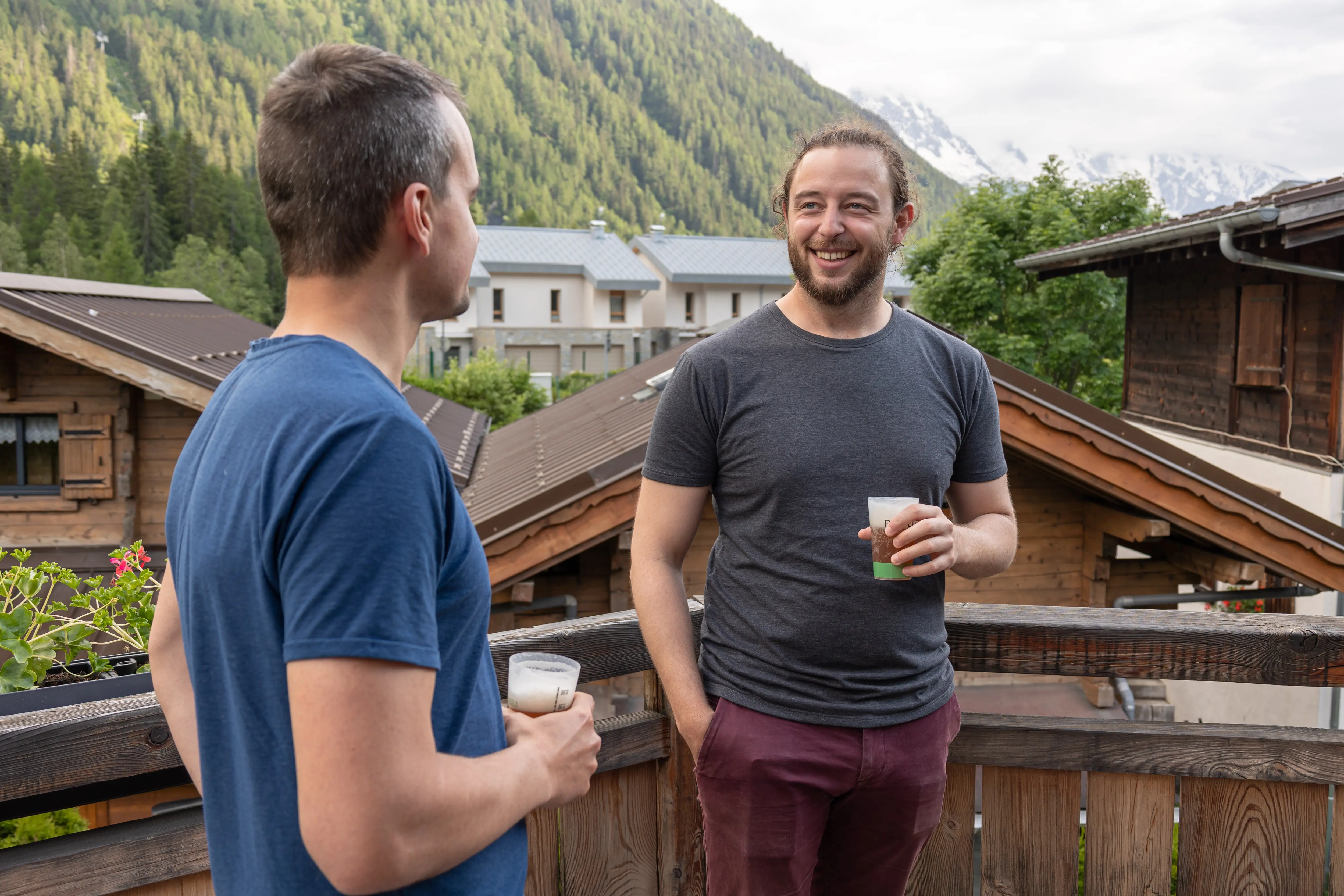 Quatre hommes debout dehors sur un chemin de gravier devant une maison en bois, jouant à la pétanque.