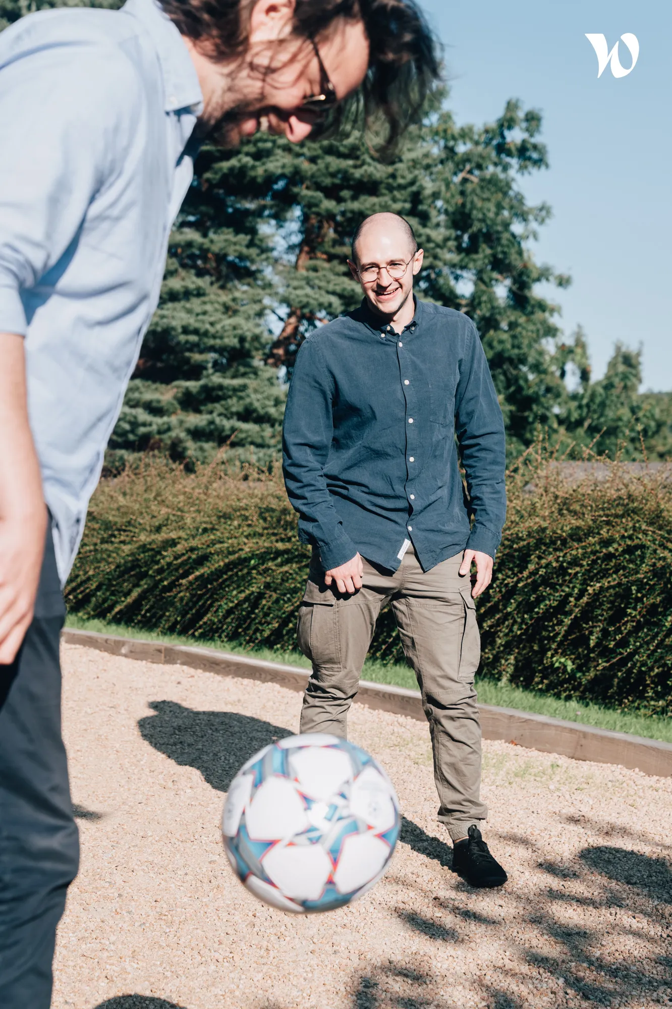 Deux hommes souriants jouant au football dans un espace extérieur avec des arbres en arrière-plan.
