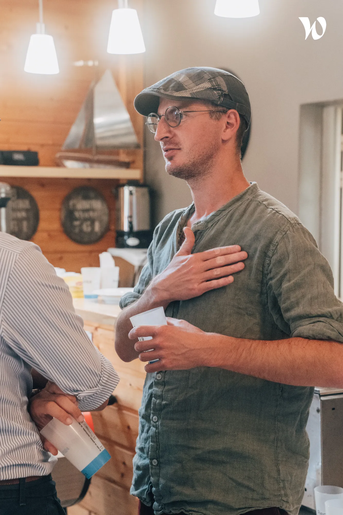 Un homme portant une casquette et des lunettes discute en plaçant une main sur sa poitrine, tenant un gobelet en plastique dans l'autre main, dans un intérieur en bois éclairé par des lampes suspendues.