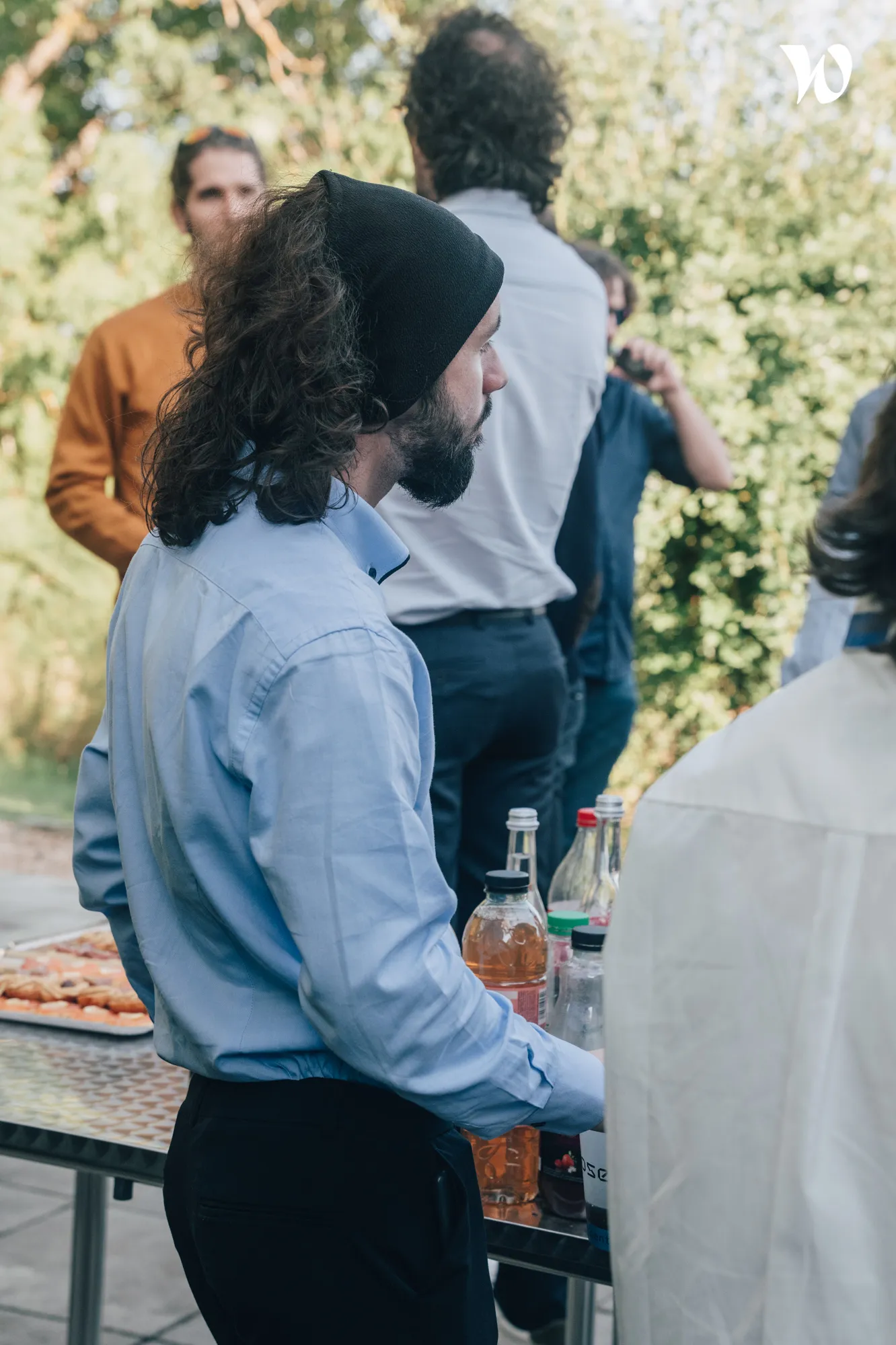 Un homme portant une chemise bleue et un bonnet noir, debout près d'une table avec des bouteilles et une plaque de nourriture en plein air lors d'un rassemblement social.