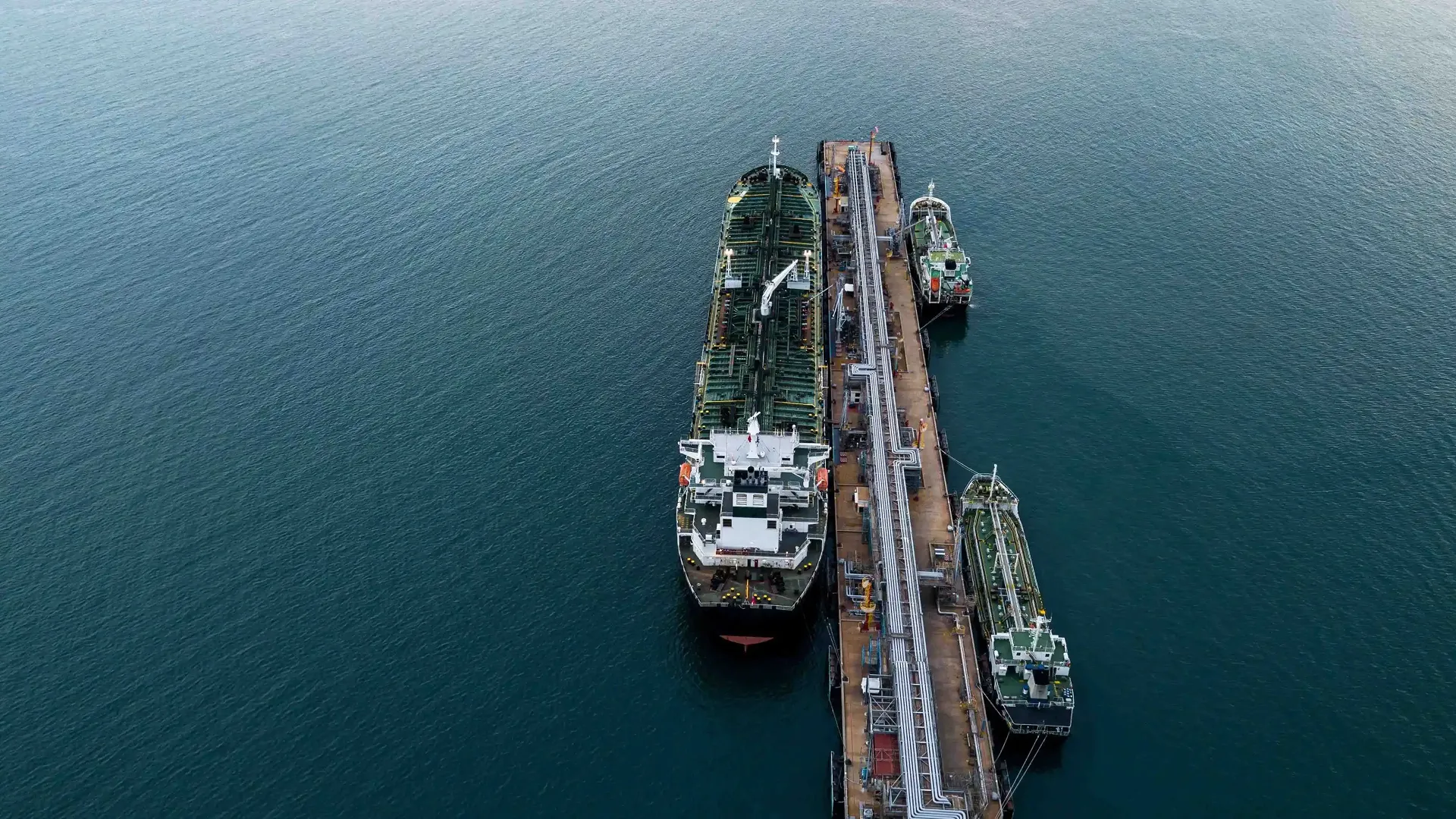 Vue aérienne d'un quai industriel avec un grand navire-citerne et deux petits bateaux amarrés sur une mer calme.