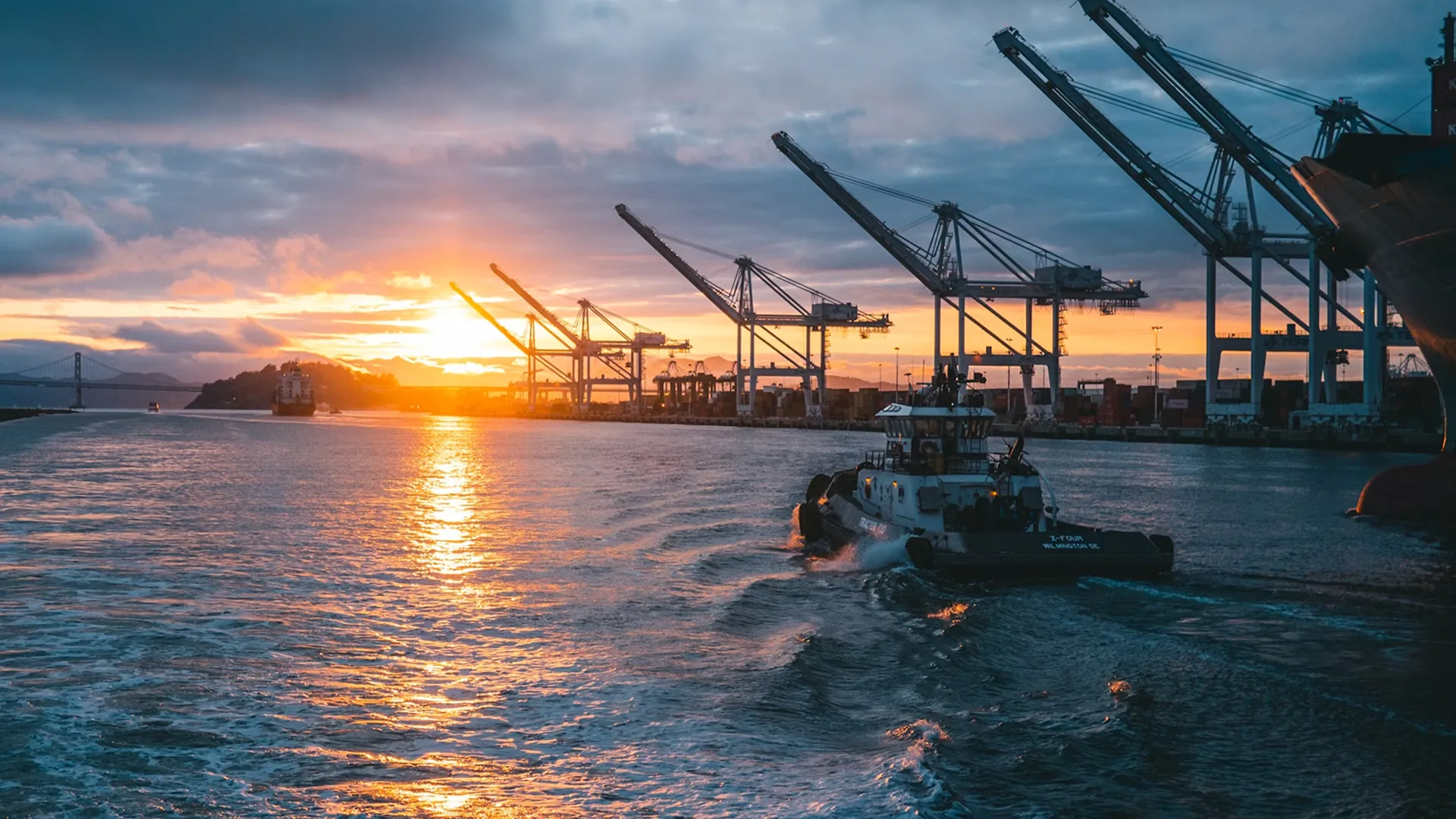 Tow boat sailing in an industrial port at sunset with several loading cranes at the dock.