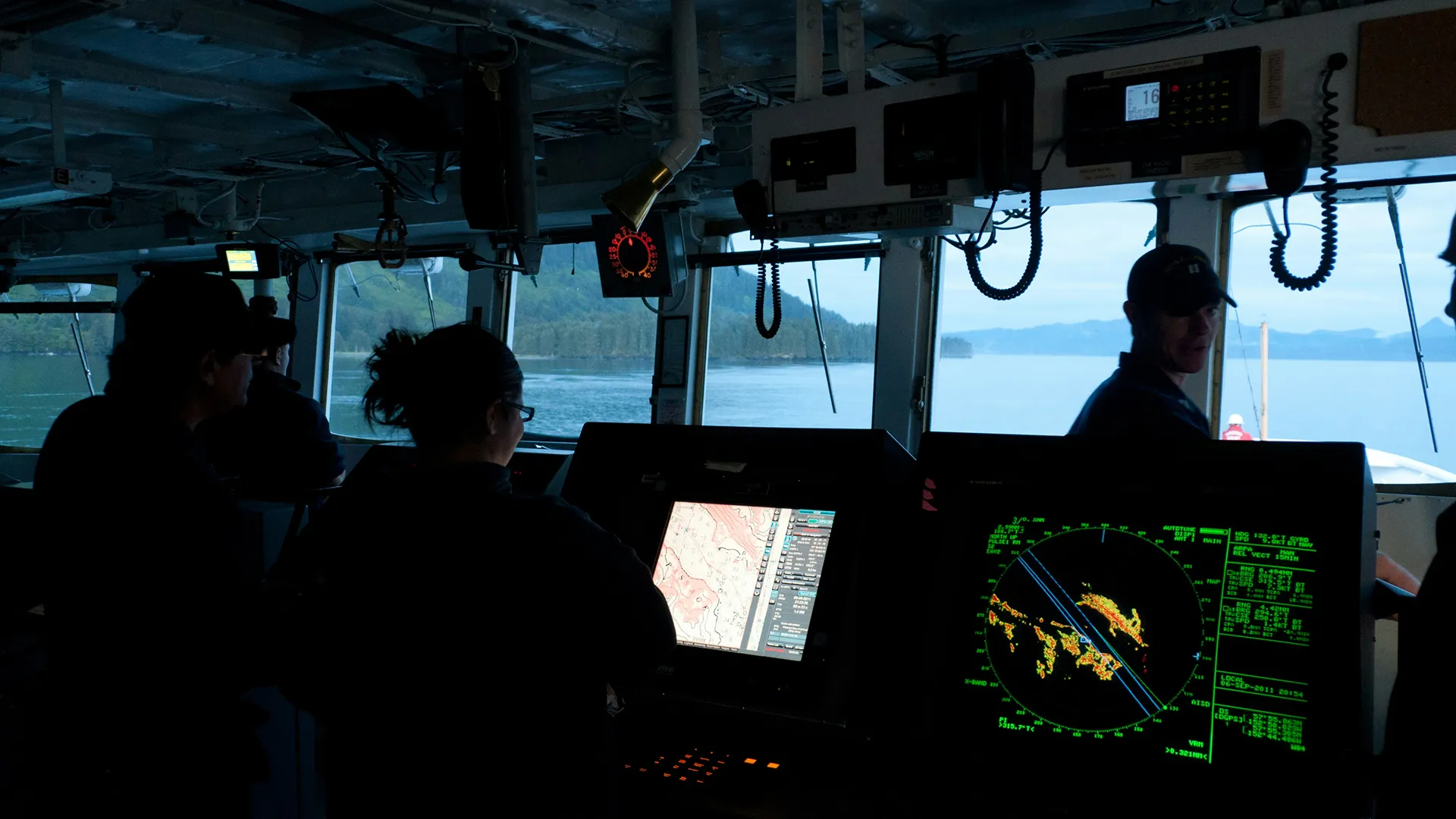 People in the cabin of a boat with navigation screens and a view of the water and the hills outside.