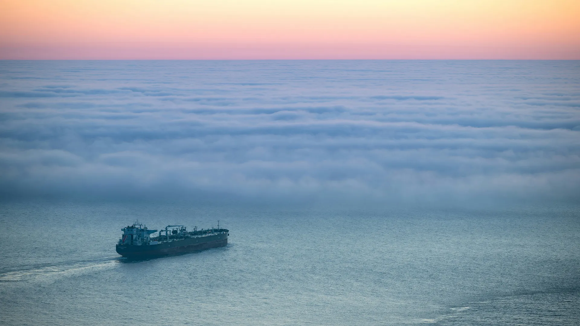 Grand cargo naviguant sur une mer calme sous un ciel aux teintes roses au coucher du soleil avec une couverture de nuages bas à l'horizon.