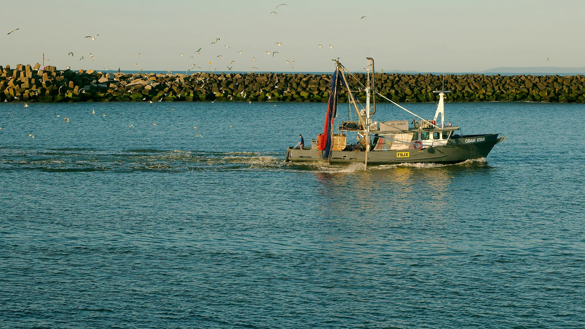 Fishing boat named Oban Star sailing through the water with seagulls flying above and a rock dike in the background.