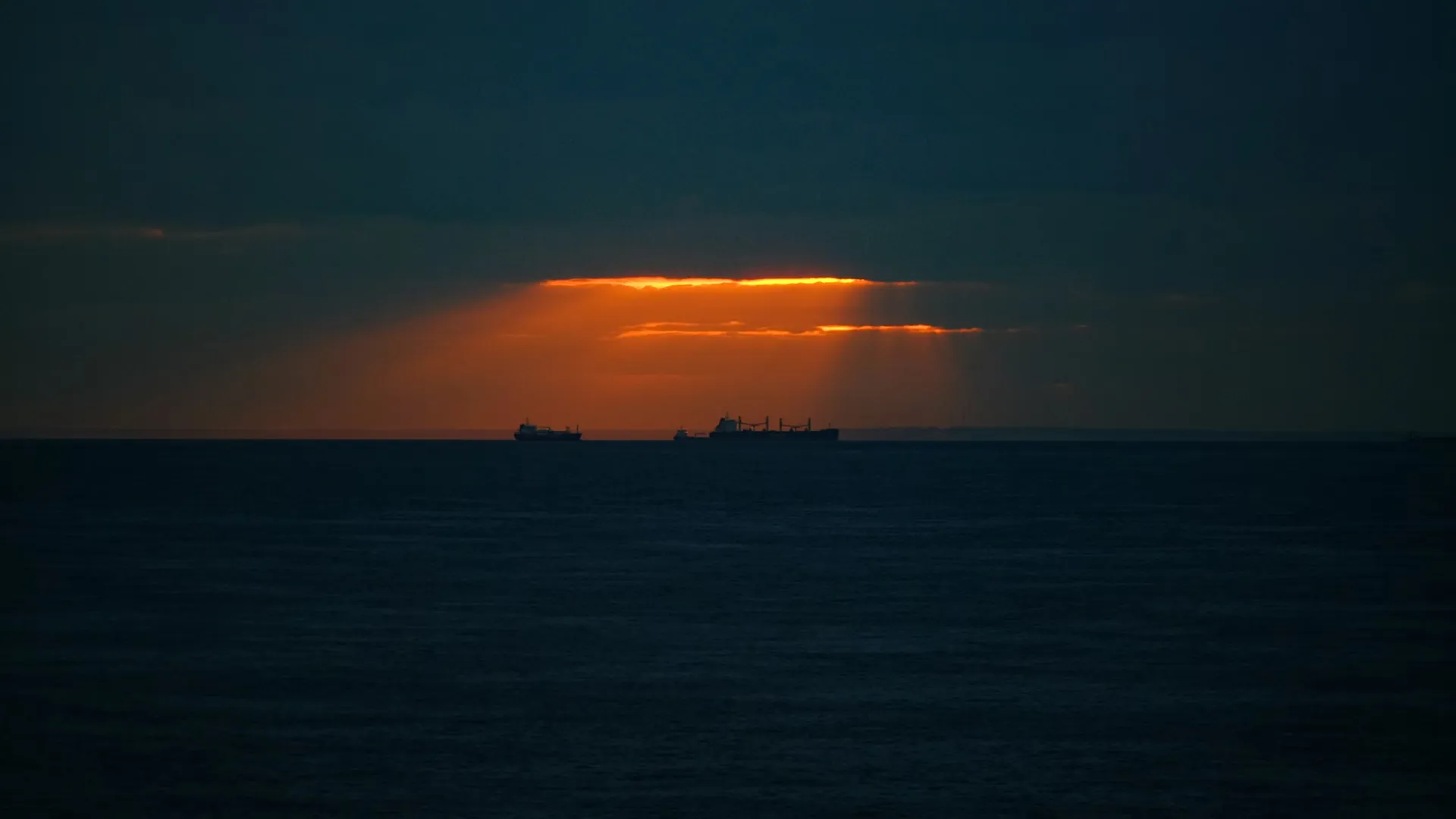Dark sky over the sea with orange sunrays piercing the clouds and three cargo ships on the horizon.