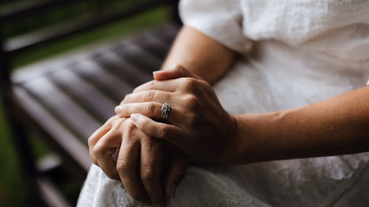 Close-up of a person’s hands resting on their lap, with a silver ring on the left ring finger, seated on a wooden bench.
