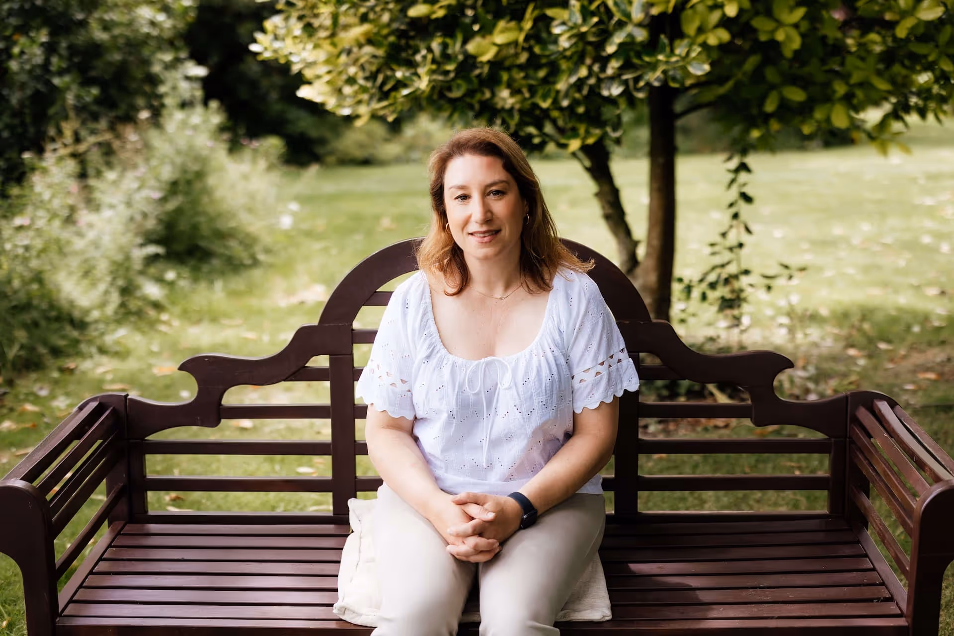 Woman with light brown hair sitting on a wooden bench in a green park, wearing a white blouse and beige pants.