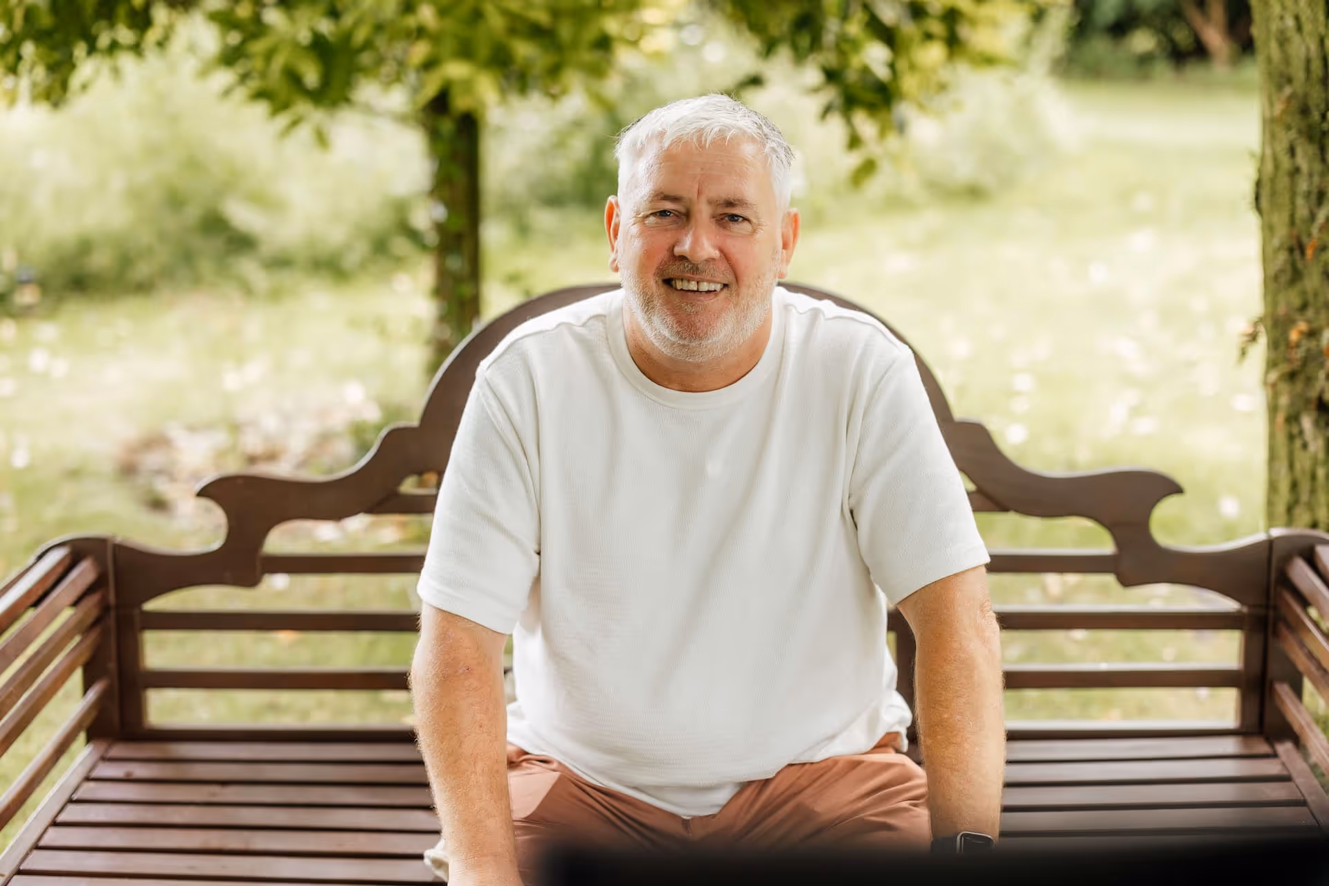 Smiling older man with white hair and beard sitting on a wooden bench outdoors.